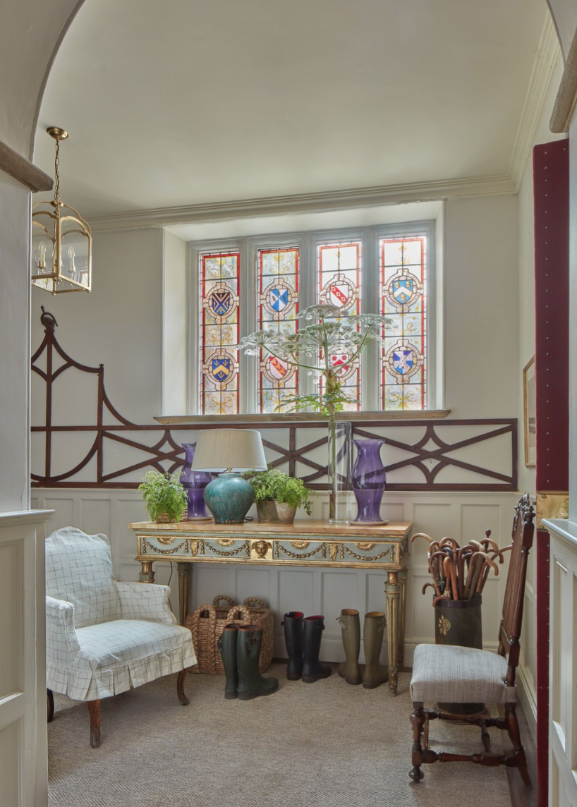 A cozy interior with a stained glass window, a vintage console table with vases and a lamp, a white armchair, a wooden chair, and rain boots on the floor.