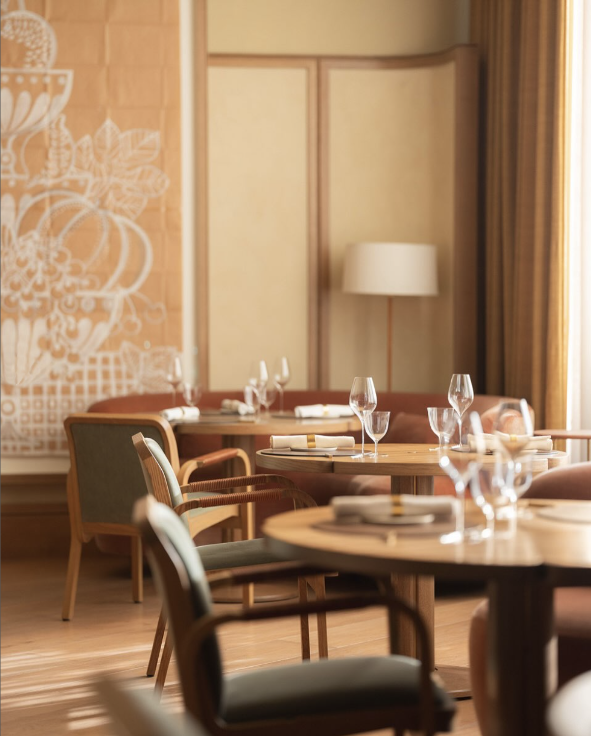 Restaurant dining area with tables set with plates, wine glasses, and napkins, featuring wooden chairs, a large wall art piece, a floor lamp, and curtains.