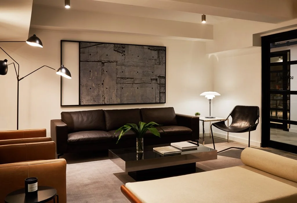 Modern living room with brown leather sofa, black armchair, glass coffee table, floor lamp, wall art, and a sliding door.
