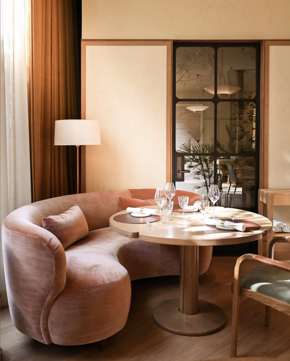 A cozy dining nook with a pink velvet curved banquette, a round wooden table set with glassware, and a standing lamp next to a beige wall with curtains. A glass door reflects plants inside.