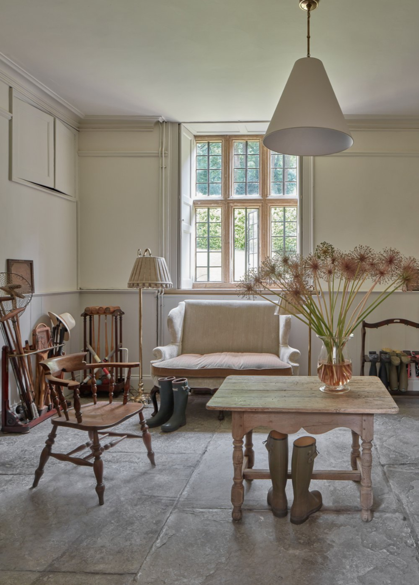 Cozy vintage living room with a large window, wooden chairs, a small table with a vase of flowers, a white sofa, and rubber boots inside.