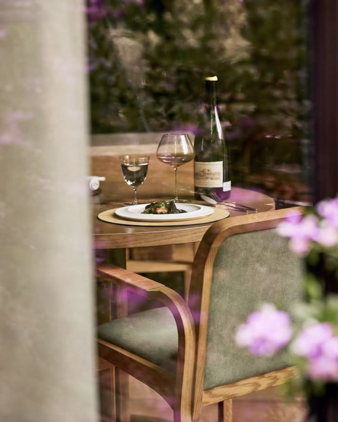 A table set for two with a wine glass, a water glass, a plate of food, and a wine bottle, seen through a window with pink flowers in the foreground.