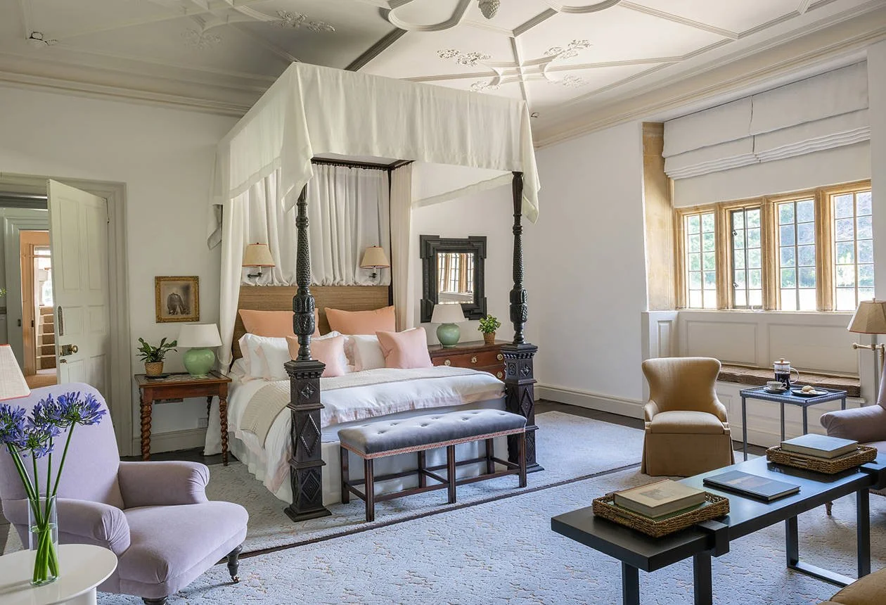 A cozy bedroom with a four-poster bed with white drapes and pink pillows, a purple armchair with purple flowers on a white side table, a beige armchair, and a black coffee table with books, in a room with large windows and ornate white ceiling details.
