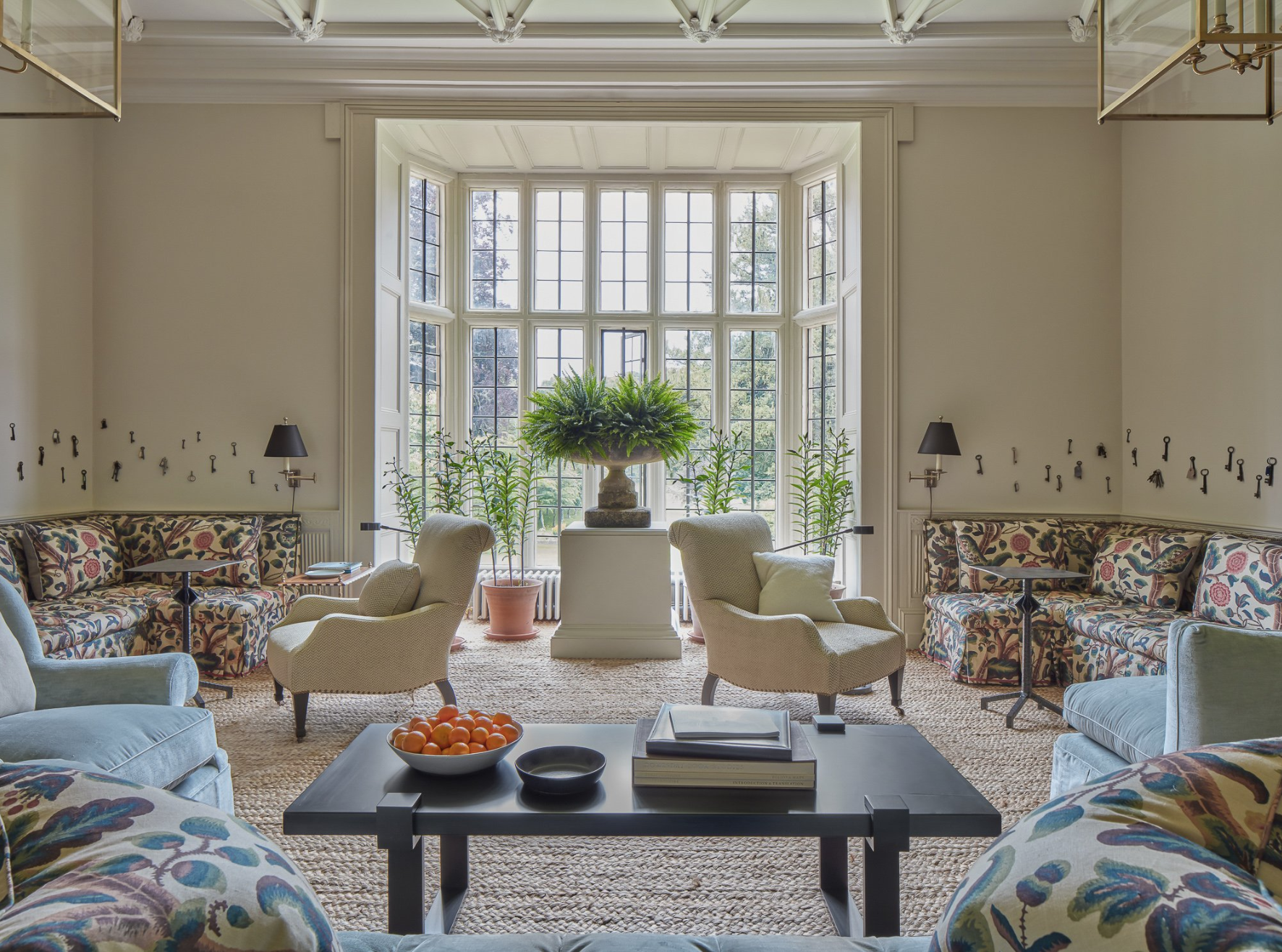 Living room with large bay window, floral-patterned sofas, armchairs, potted plants, and a coffee table with bowls of oranges and black bowls.