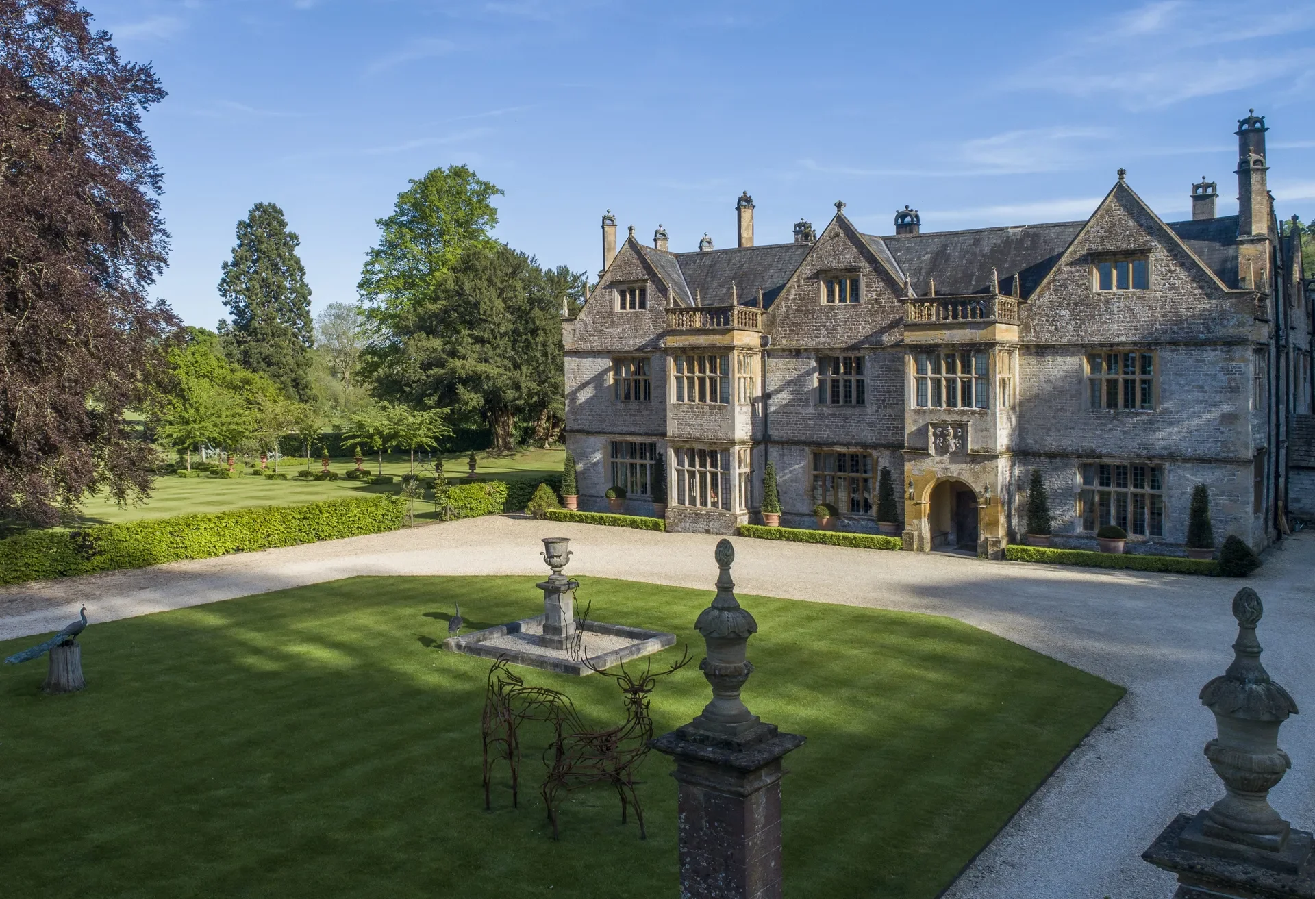 A large historic stone mansion with multiple chimneys and gabled roofs, surrounded by manicured lawns, trees, and a gravel courtyard in front, under a clear blue sky.