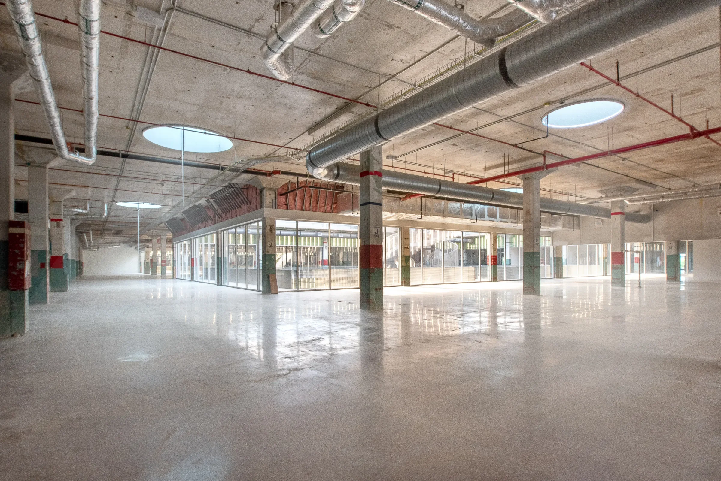 An empty indoor industrial space or parking garage with concrete floors, ceiling, and supporting columns. Exposed ducts, pipes, and circular skylights are visible on the ceiling.