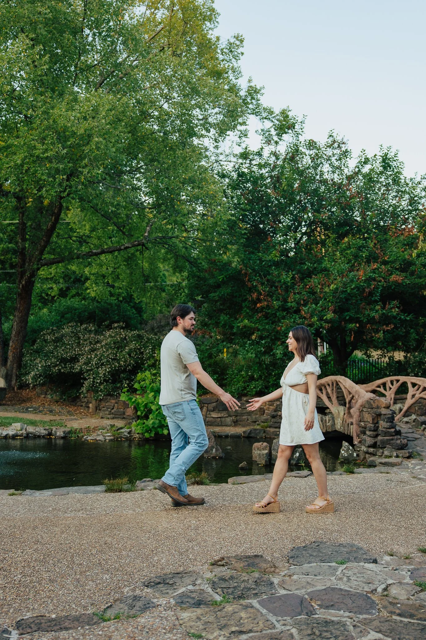 A man and a woman are standing on a stone pathway by a small body of water in a park, holding hands and facing each other, surrounded by green trees.