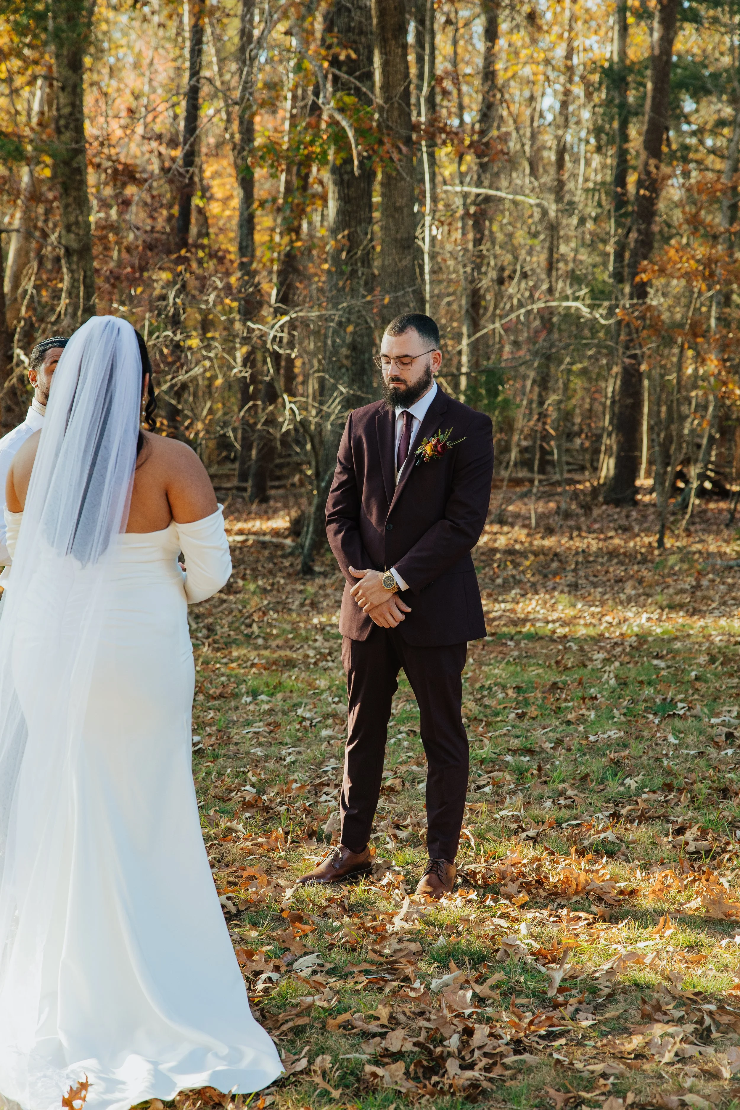 A bride and groom standing outdoors in a wooded area with autumn leaves on the ground. The bride, with a long white dress and veil, faces away from the camera, while the groom, in a dark suit with a boutonniere, faces downward with a serious expressi
