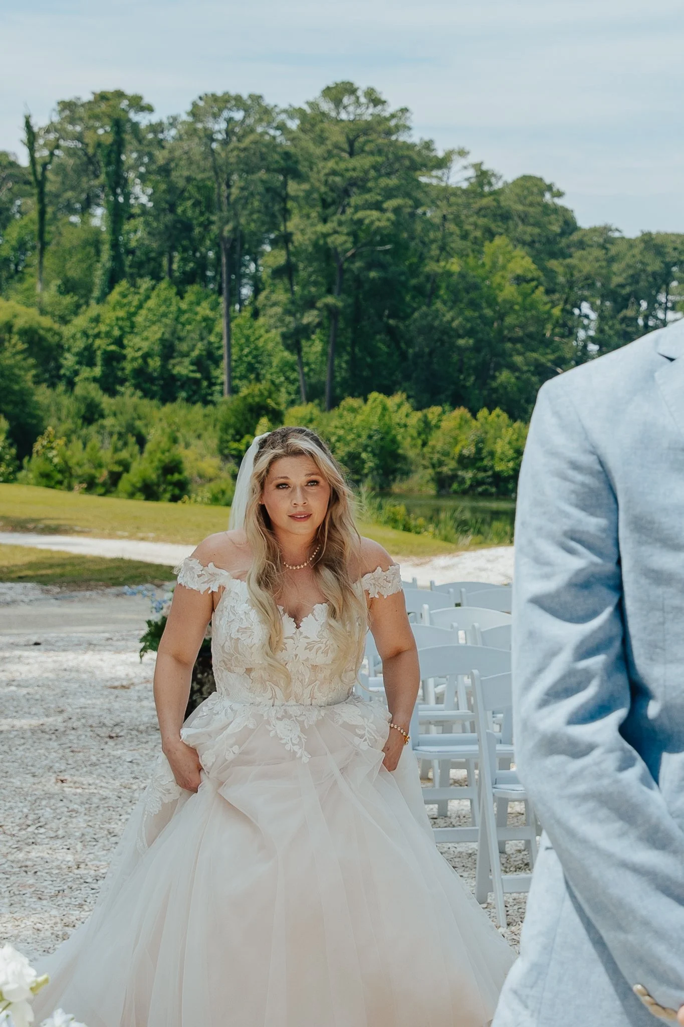 A bride with long blonde hair wearing a white strapless wedding dress with lace details, standing outdoors on a gravel pathway, with rows of white chairs behind her, trees in the background, and a person in a light-colored suit partially visible on t