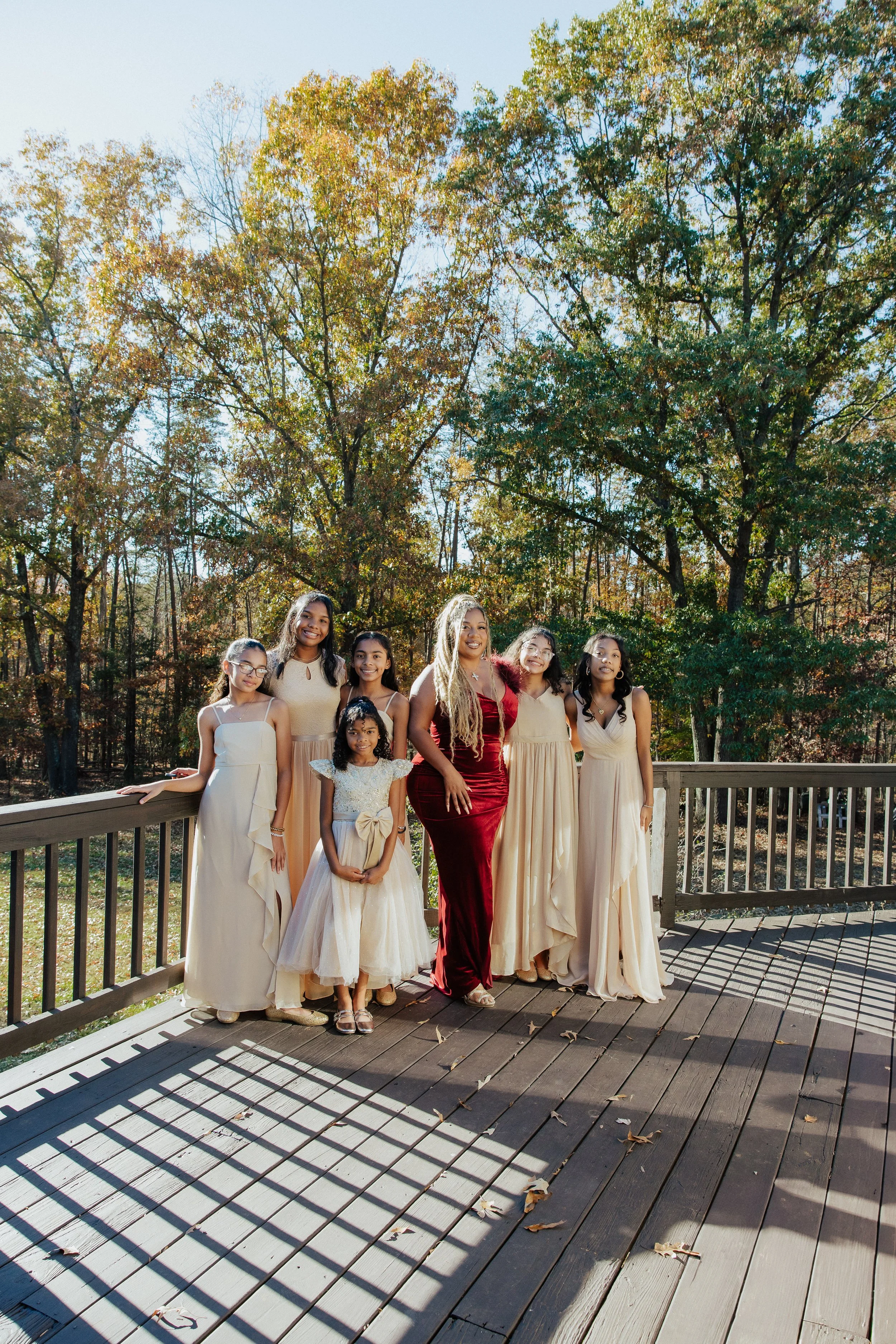 Group of women and girls standing on a wooden deck outdoors with fall trees in the background, some women wearing elegant dresses, one in a red dress, others in beige or cream dresses, smiling.