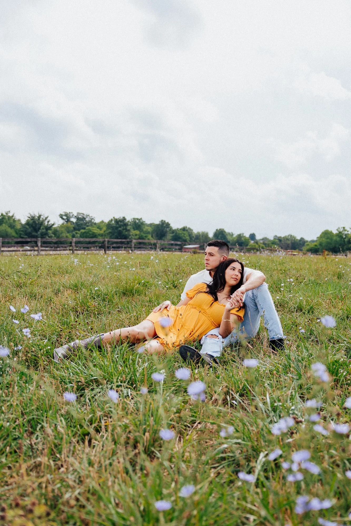A young man and woman sitting in a field of grass and small purple flowers, with a wooden fence and trees in the background, under a cloudy sky.