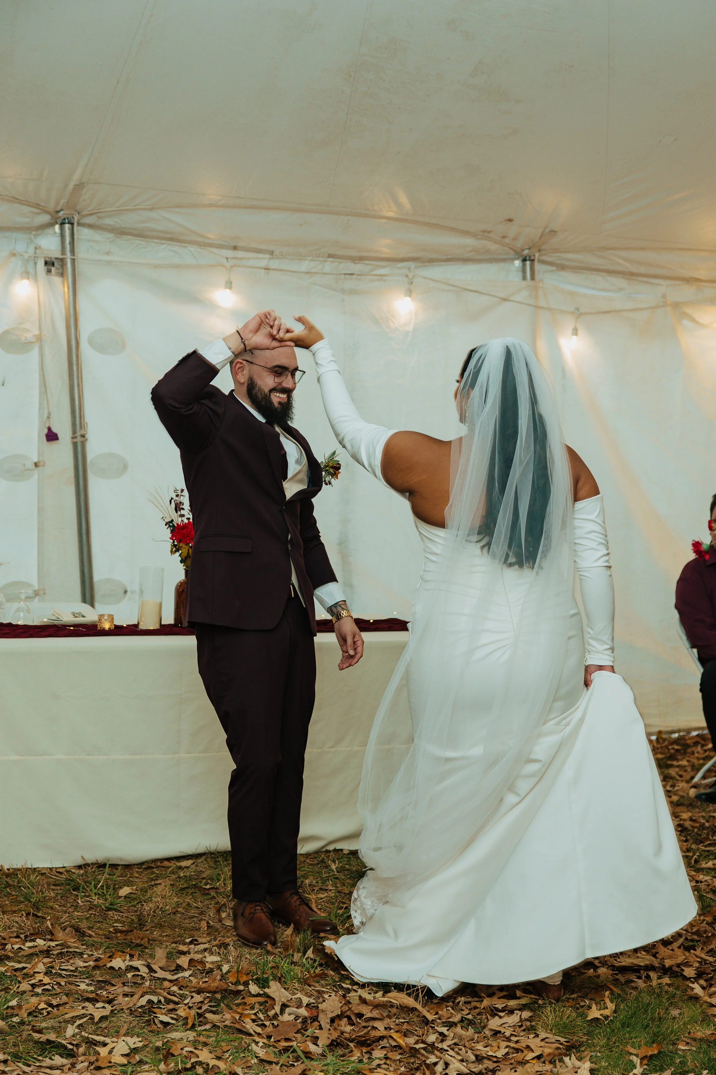 A newlywed couple dancing at their outdoor wedding reception, under a white tent decorated with string lights. The bride is wearing a long white wedding gown and veil, and the groom is in a dark suit with a boutonniere. They are smiling and holding h