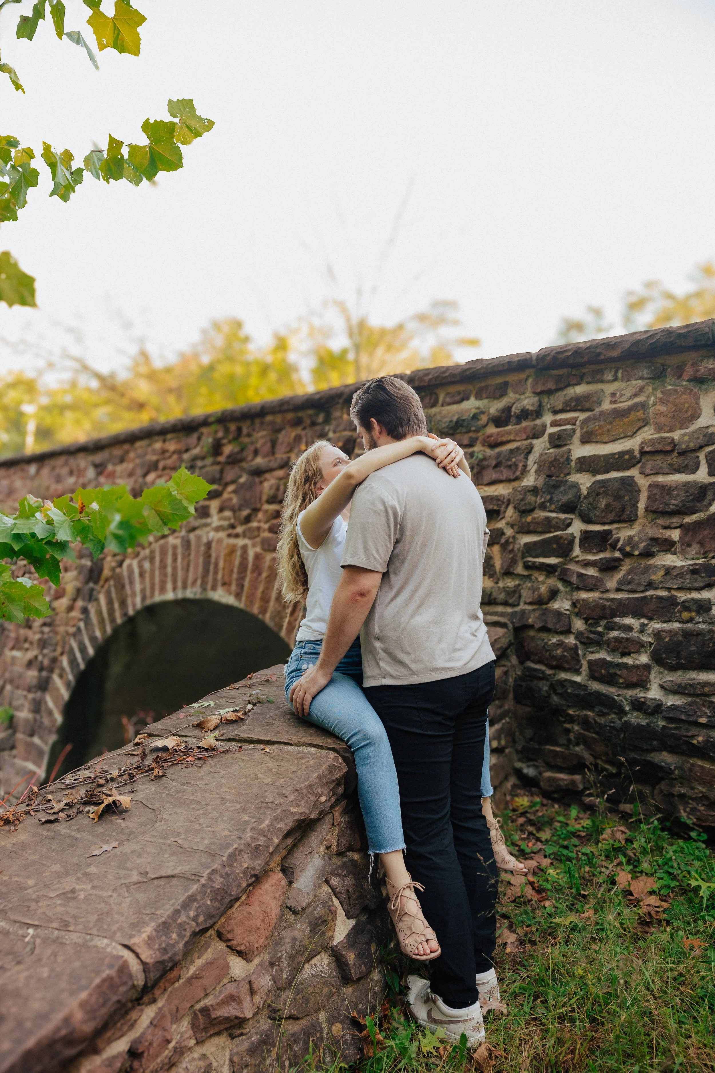 A couple is embracing on a stone ledge next to a brick arched bridge, with autumn trees and green leaves in the background.
