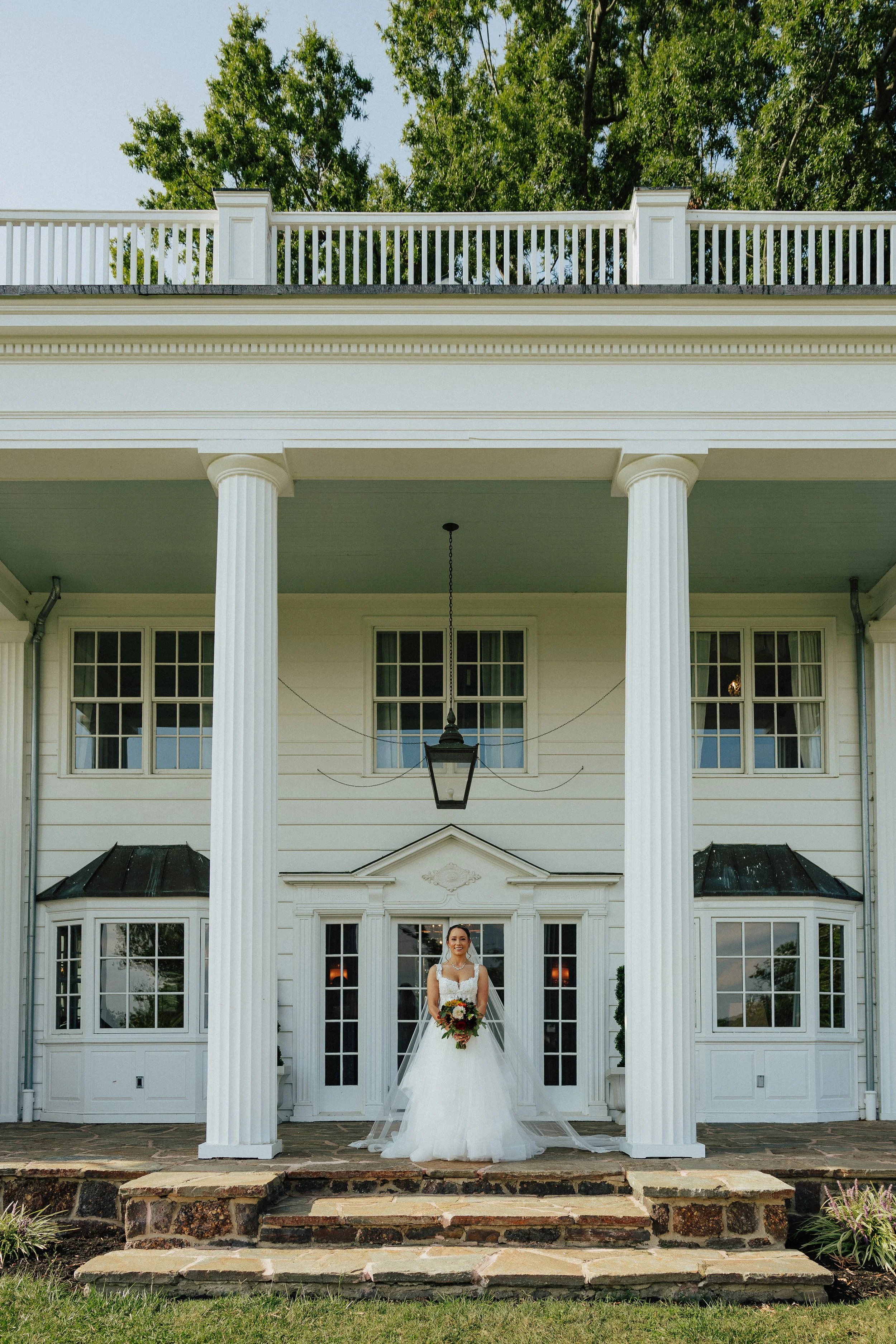 A bride in a white wedding gown holding a bouquet of flowers standing on stone steps in front of a white house with tall columns and a porch. The house has multiple windows and a lantern hanging from the porch ceiling. Trees are visible behind the ho