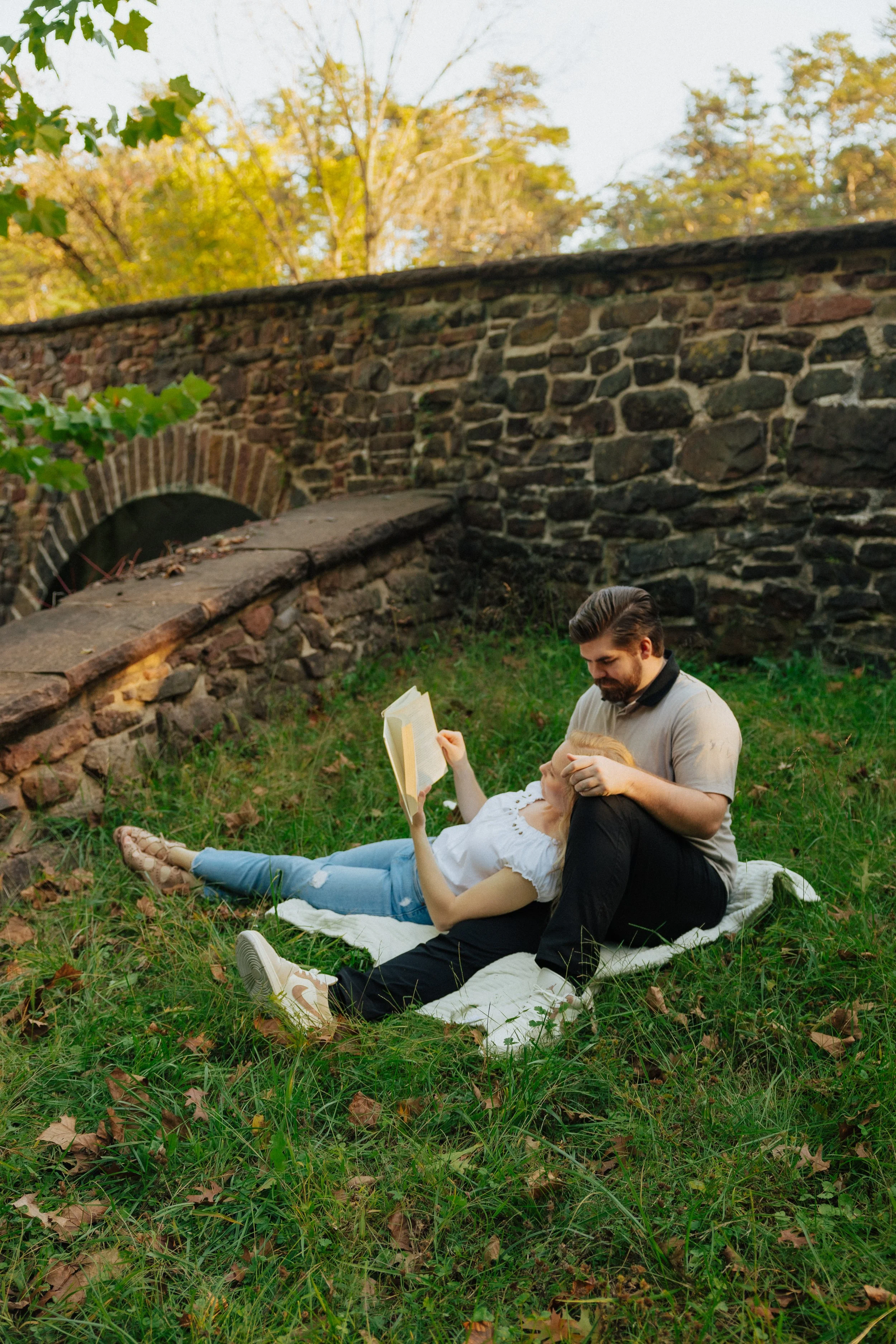 A man and woman relaxing outdoors on a blanket in a grassy area with fallen leaves, reading books with a stone bridge and autumn trees in the background.