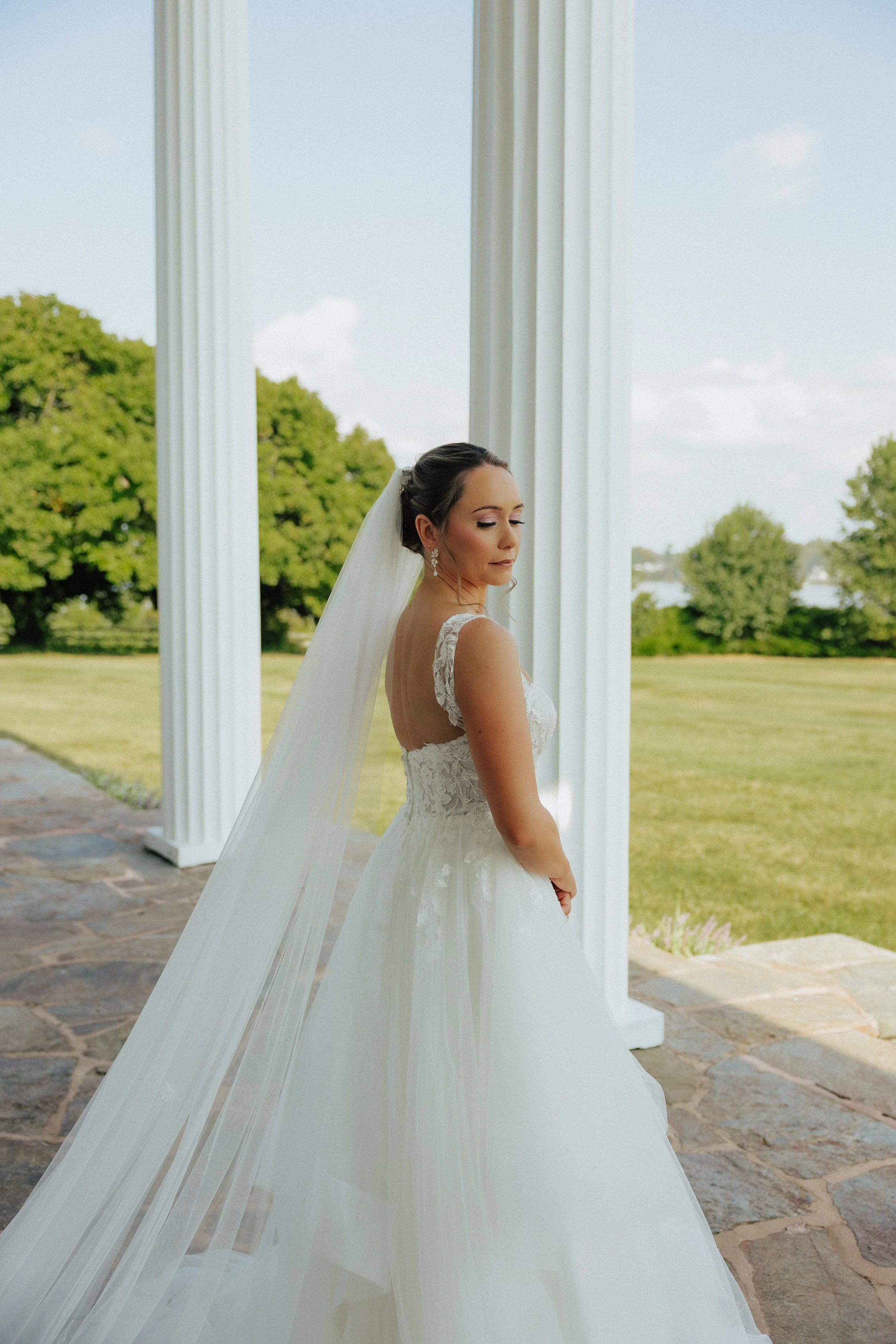 A bride in a white wedding dress standing outdoors under large white columns with green trees and a blue sky in the background.