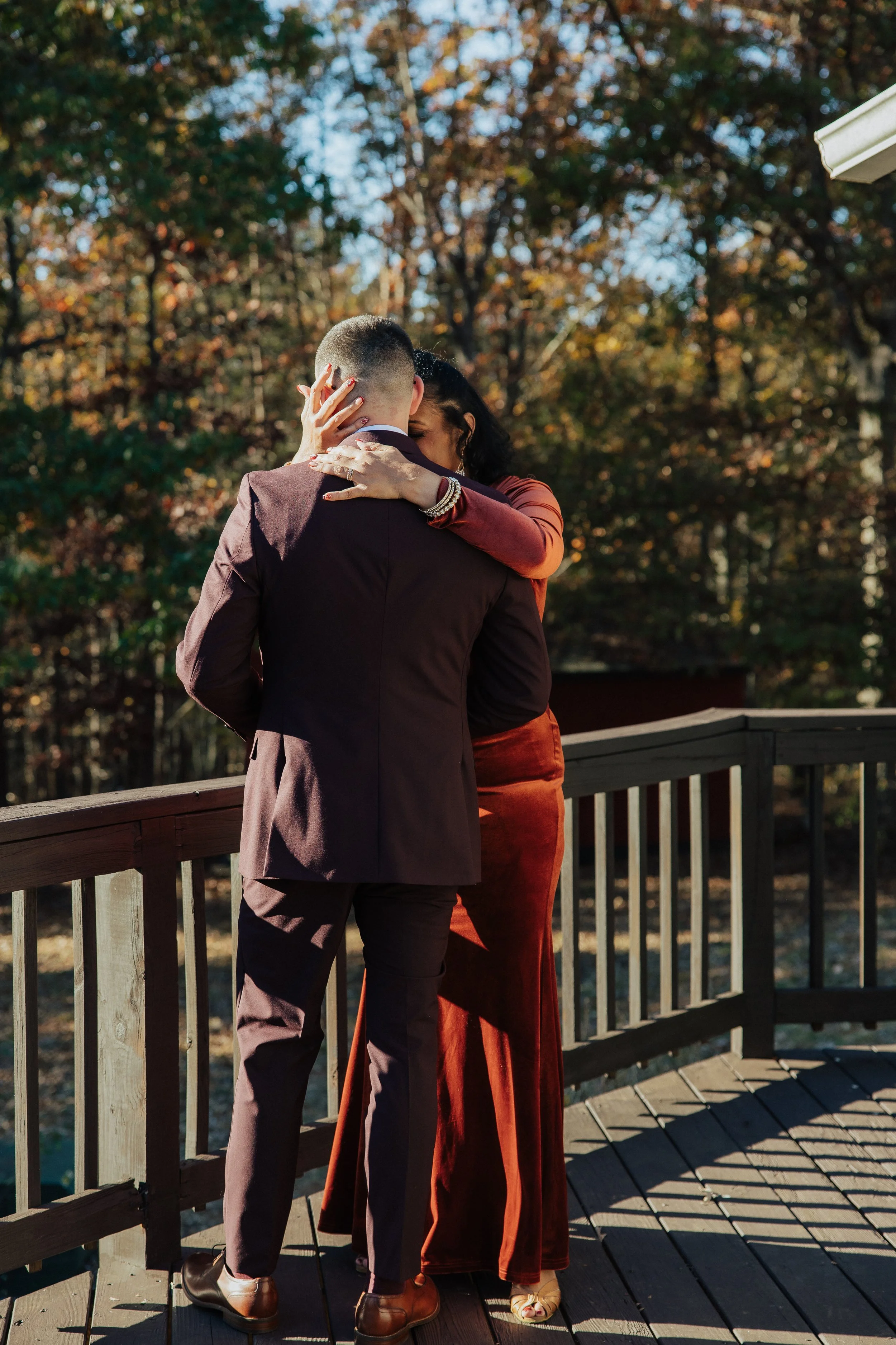 A couple hugging on a wooden deck outdoors, with trees in the background, during autumn.