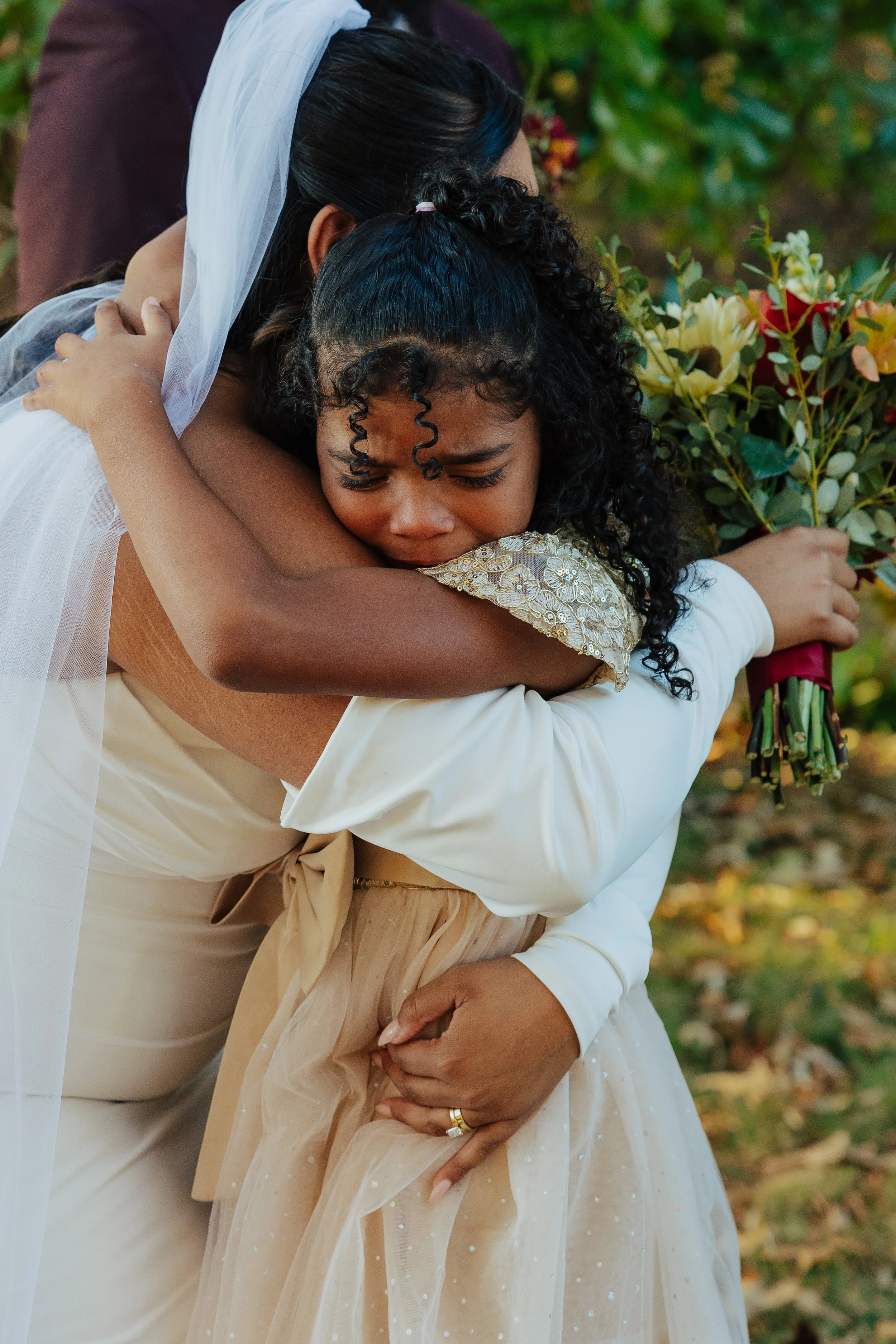 Two women hugging, one woman wearing a white dress with golden embroidered sleeves and the other in a beige dress with a sheer, dotted overlay, outdoors with fallen leaves and green foliage.