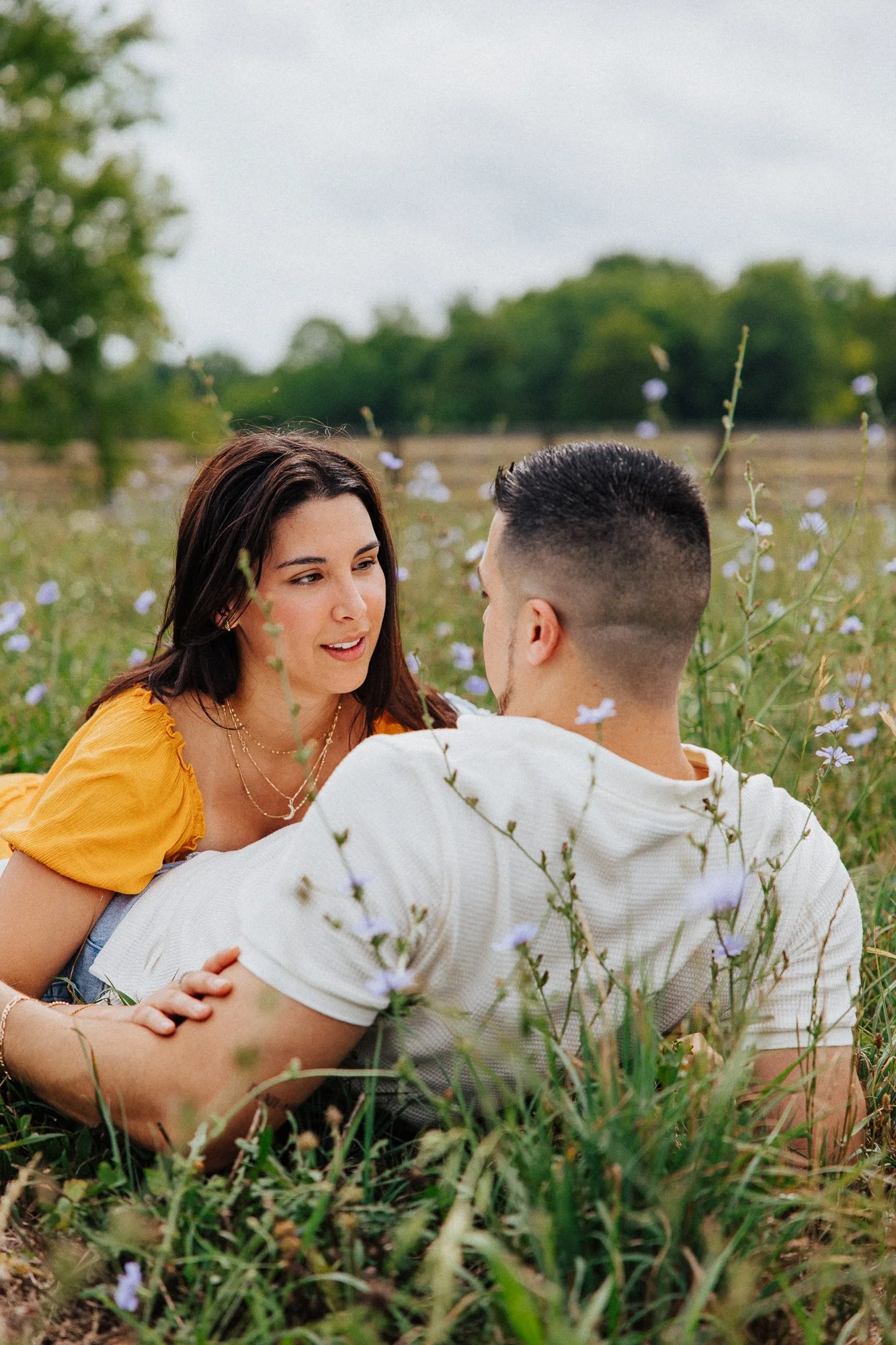 A woman and a man sitting on the grass in a field of small purple flowers, facing each other and engaged in conversation, with trees and a cloudy sky in the background.