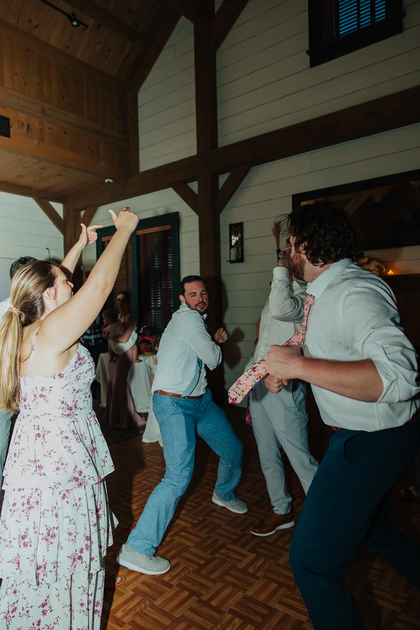 People dancing energetically at a social gathering or party in a rustic indoor space with wooden beams and paneling, some with their hands raised, enjoying the music.