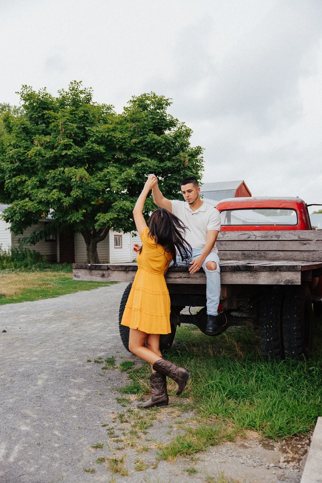 A young woman with long dark hair in a yellow dress and cowboy boots dancing with a young man in a white t-shirt and ripped jeans sitting on the back of a red truck. The truck is parked outdoors near a house with a large green tree and cloudy sky in 