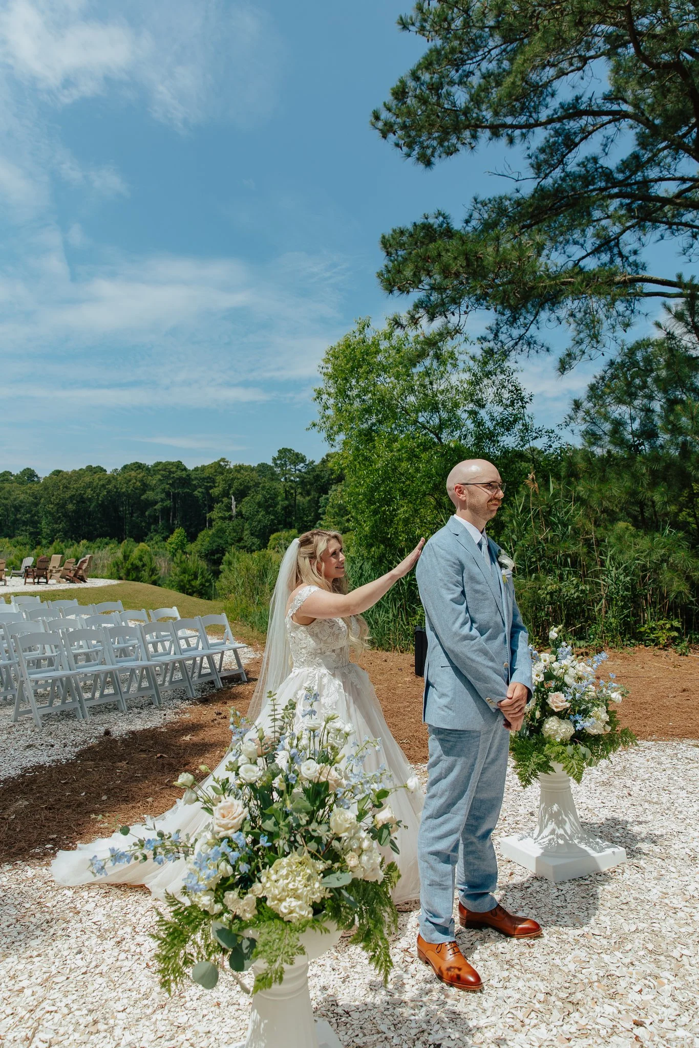 A bride and groom are standing outdoors during a wedding ceremony, with the bride reaching out to touch the groom's shoulder. The scene is decorated with floral arrangements, and chairs are set up for guests in the background. The setting is a bright
