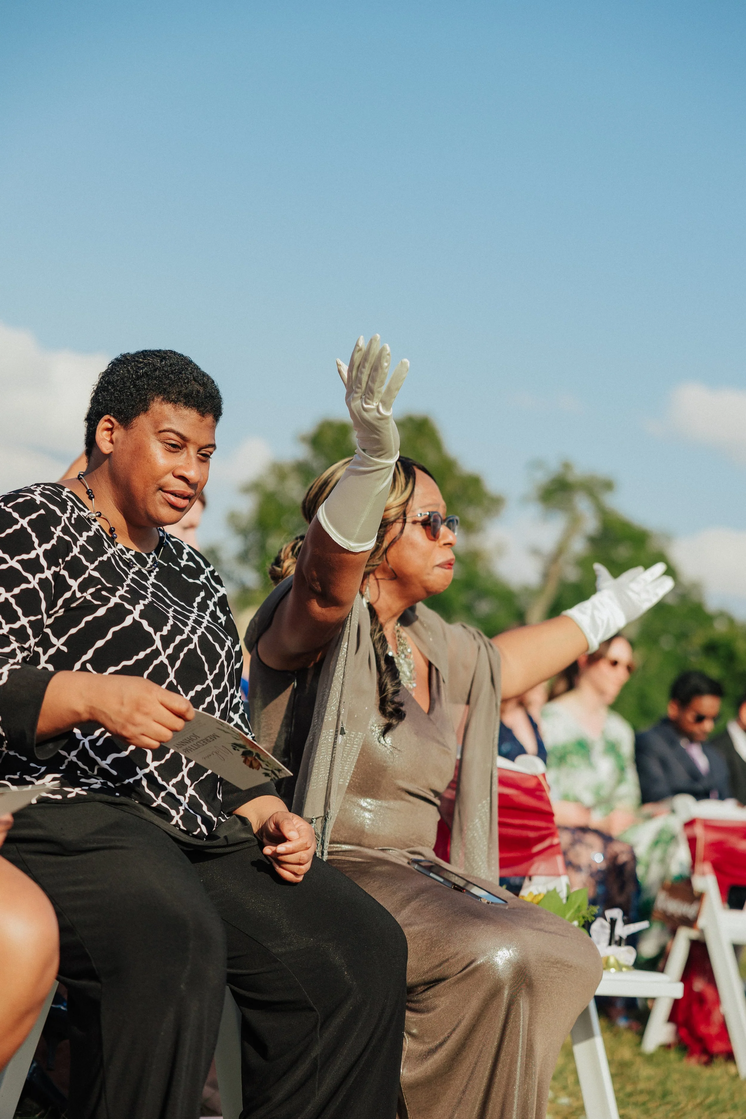 A group of women sitting outdoors during daytime, participating in a memorial or tribute event, with one woman raising her hand