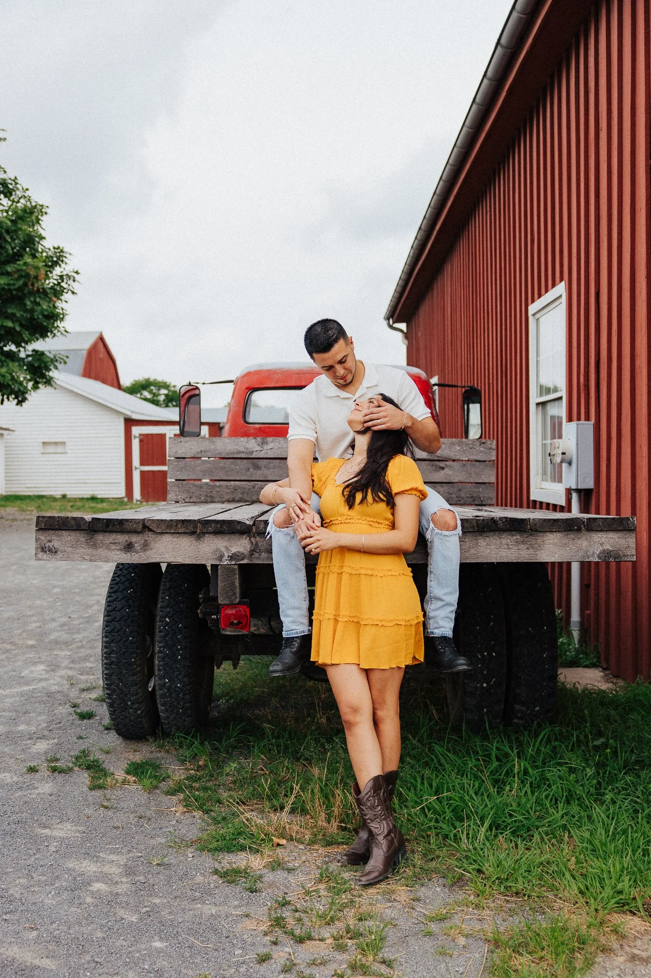 A young man and woman on a farm, with the man sitting on a truck bed and the woman standing with her back on the truck, both smiling. The woman wears a yellow dress and cowboy boots, and the man wears a white shirt and ripped jeans. There are red barns and white buildings in the background.