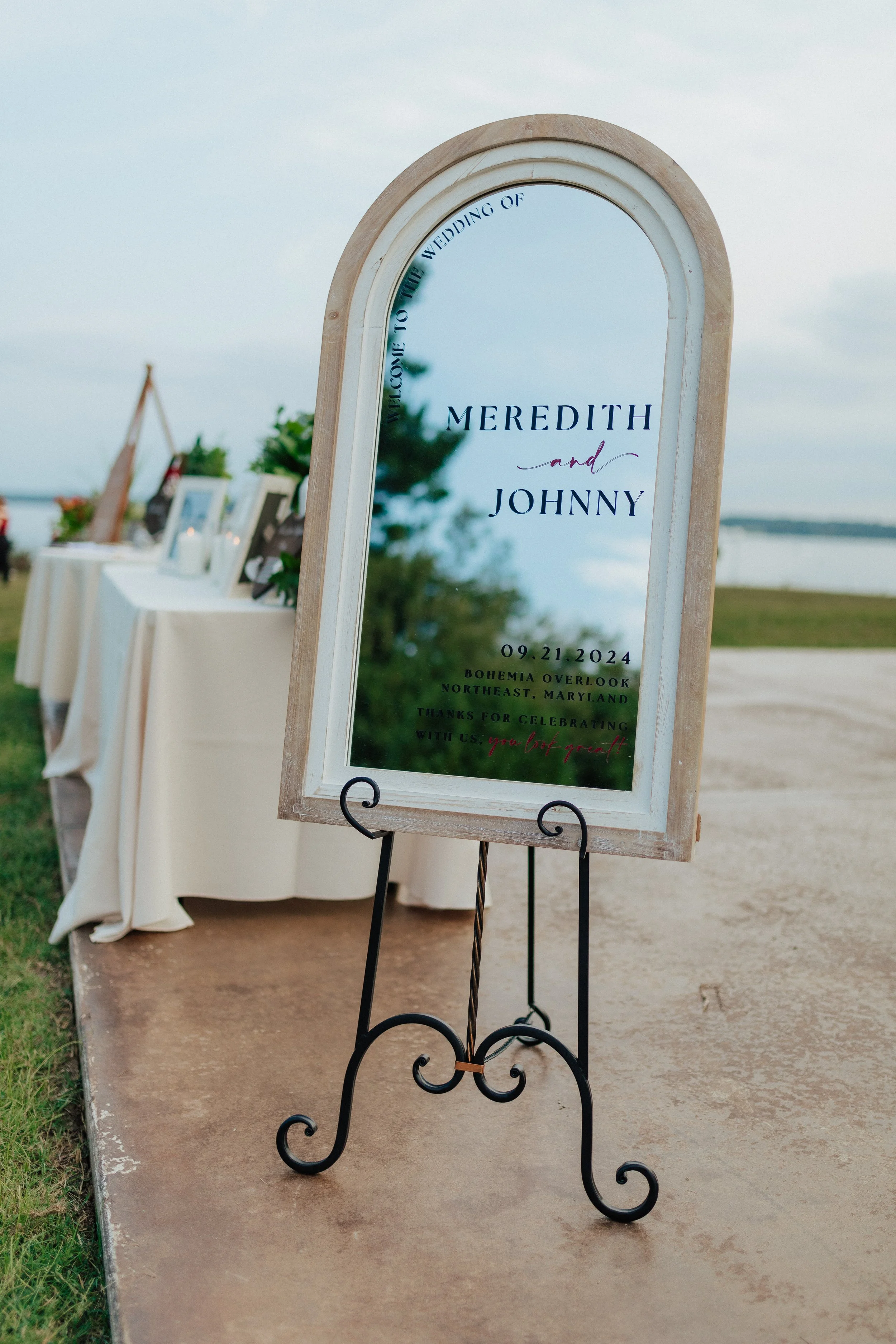 Mirror sign welcoming to a wedding of Meredith and Johnny on September 21, 2024, at Bohemia Overlook, Northeast Maryland, near a waterfront with outdoor decorations and tables with framed photos.