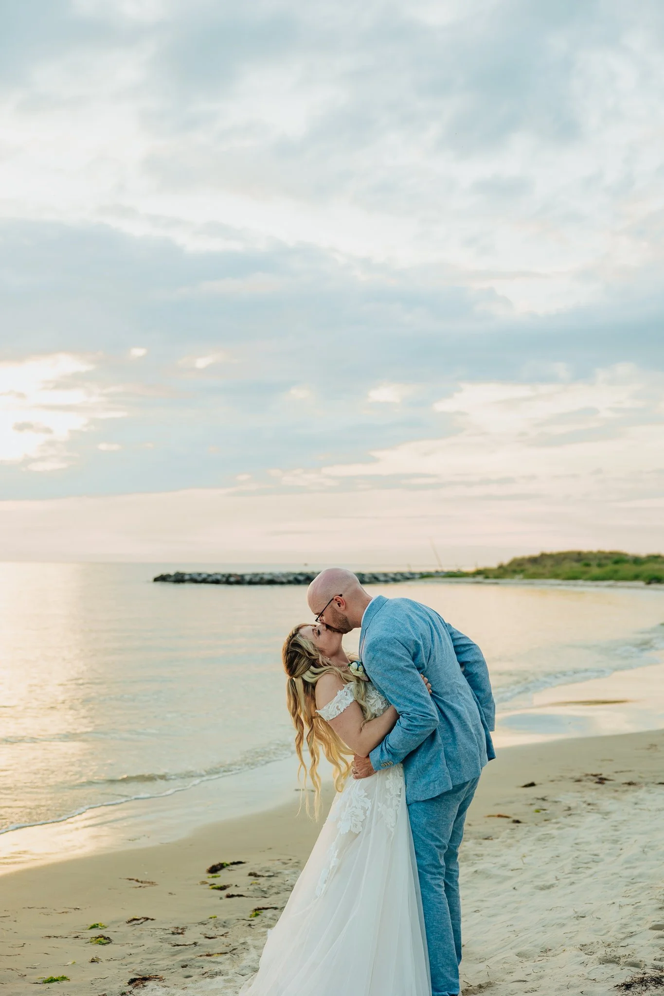 A couple sharing a kiss on the beach during sunset, with the woman in a white wedding dress and the man in a light blue suit.