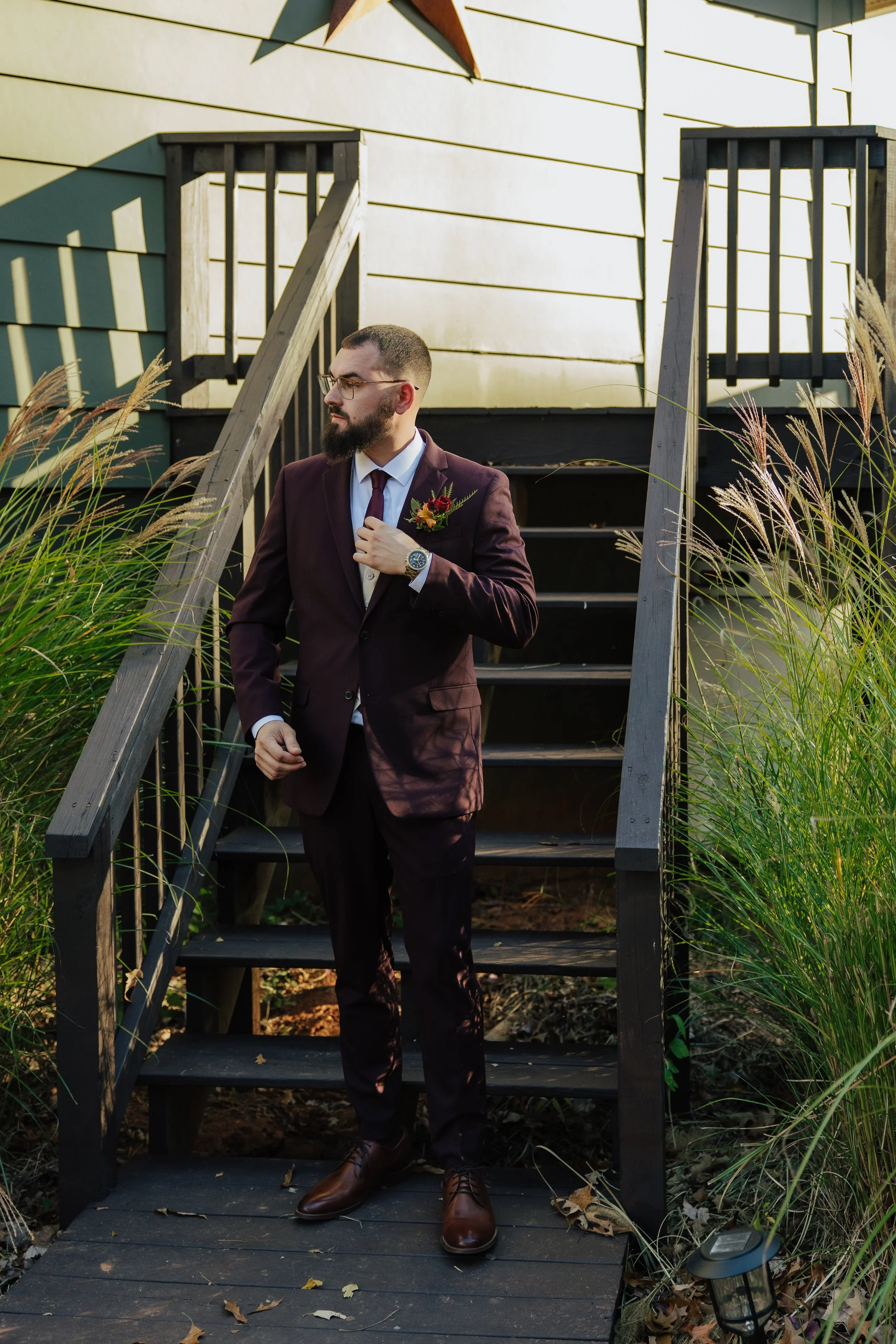 A man in a maroon suit, wearing glasses and a watch, stands on a wooden deck with stairs behind him. He has a boutonniere on his lapel, holding his jacket lapel with his right hand, looking to the side, with tall grass and a star decoration on the ho