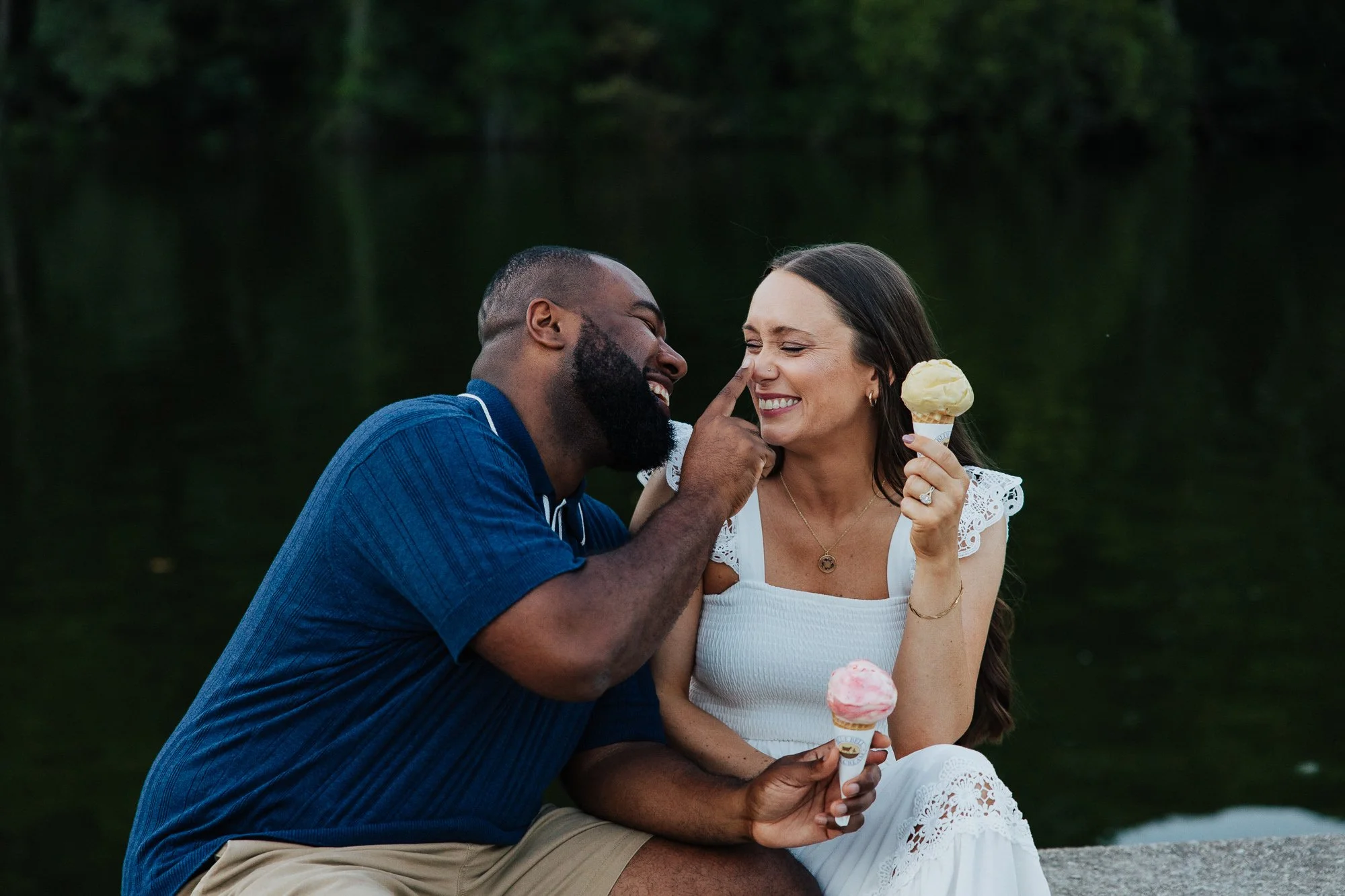 A happy black man and white woman sharing a moment, the man playfully touching the woman's nose while she smiles and holds an ice cream cone, outdoors near a body of water and trees.