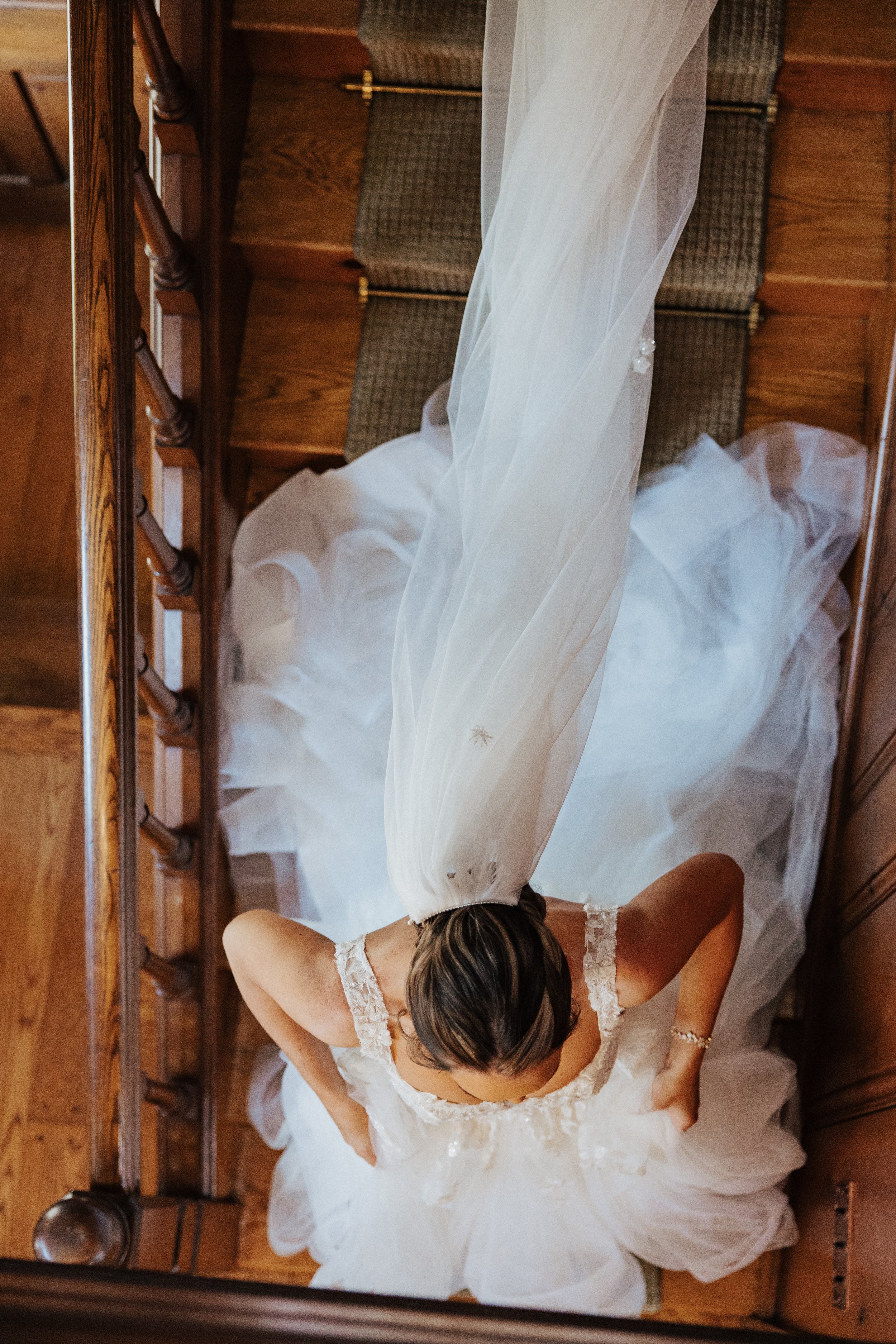 A bride with brown hair styled in waves, wearing a white wedding gown with lace straps, is getting ready in a wooden hallway or staircase area. She is holding the skirt of her dress as she prepares, with the image taken from above.