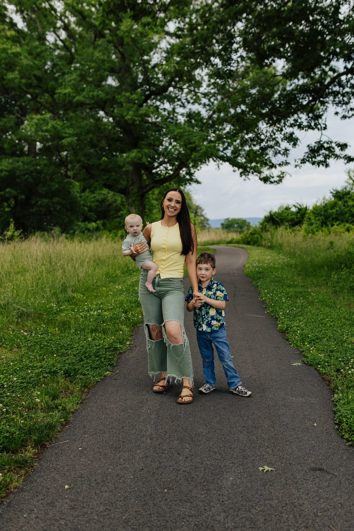 A smiling woman with long dark hair wearing a yellow sleeveless top and ripped jeans holding a young child, and a young boy in a floral shirt standing on a paved walking trail in a lush green park.