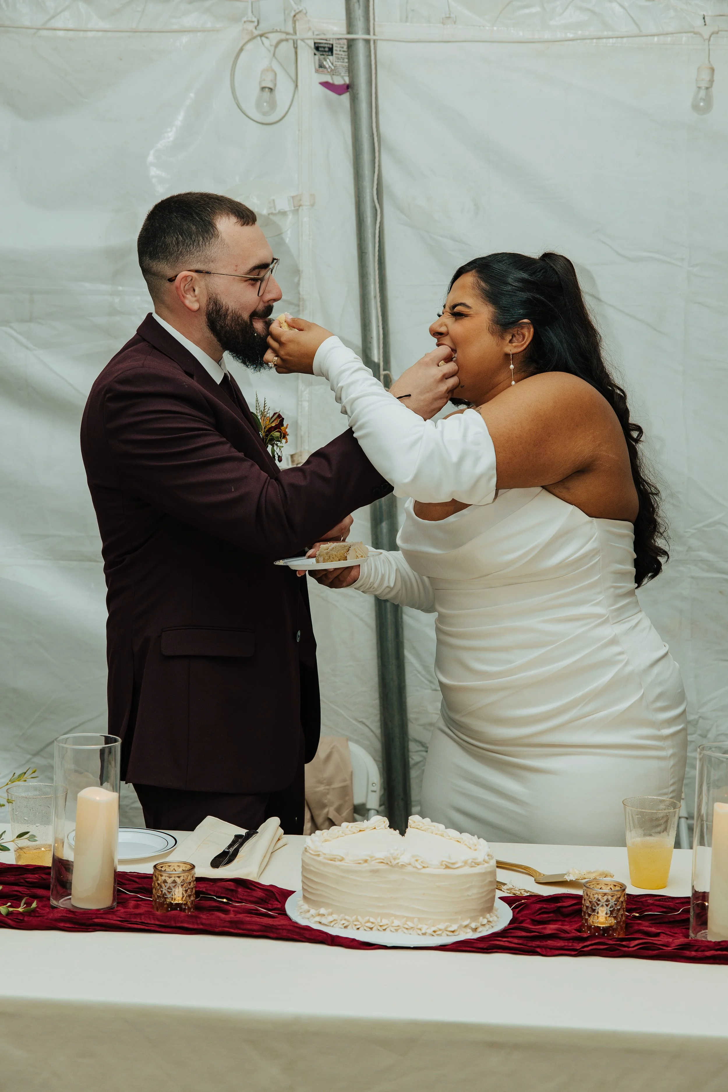 A bride and groom are feeding each other wedding cake at their reception. The table has candles, glasses, and a decorated cake on a red table runner.