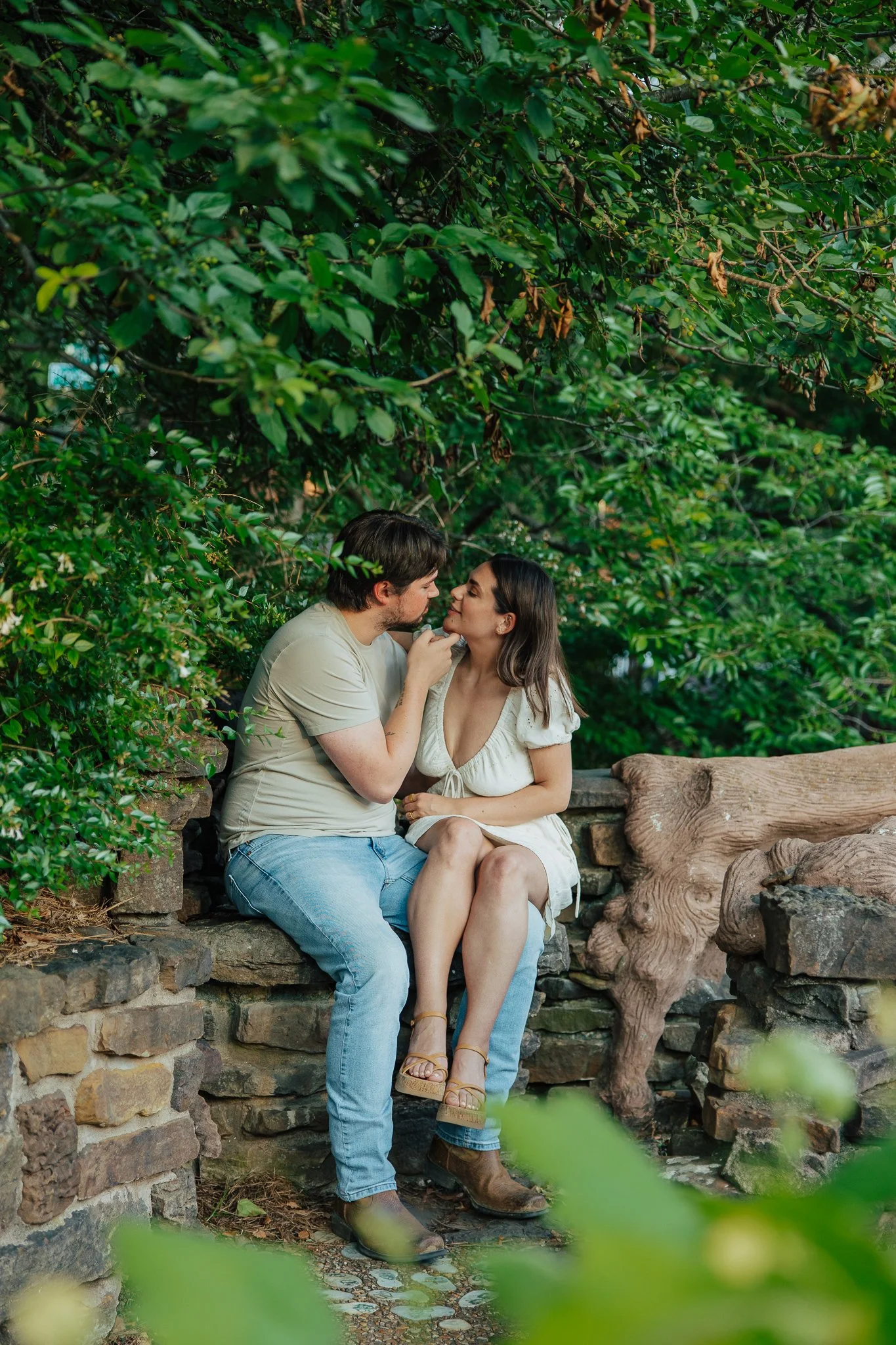 A man and woman sitting close together on a stone bench in a lush, green outdoor setting, looking into each other's eyes, with the man touching the woman's face.