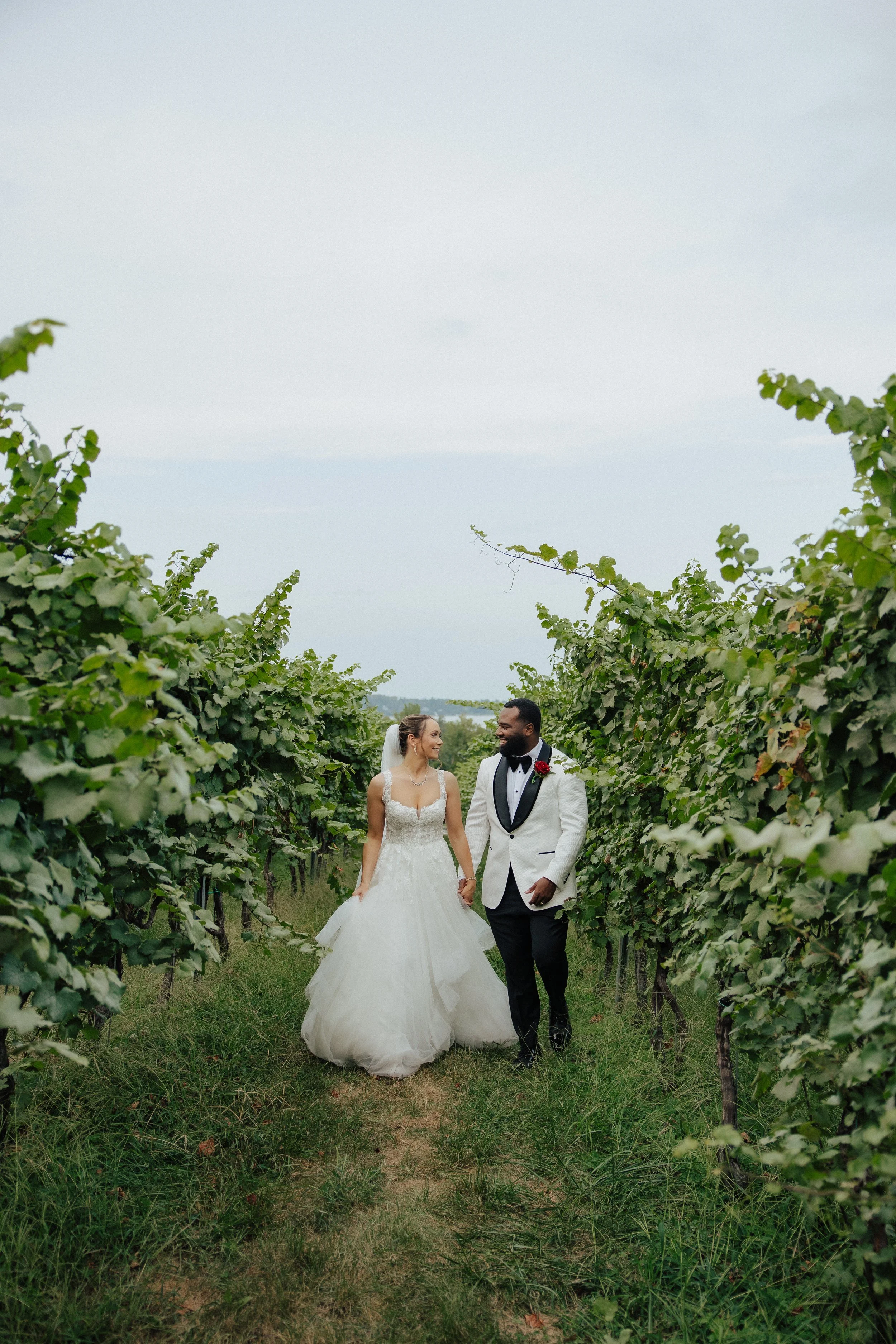 A bride and groom walking through a vineyard, holding hands and smiling at each other.