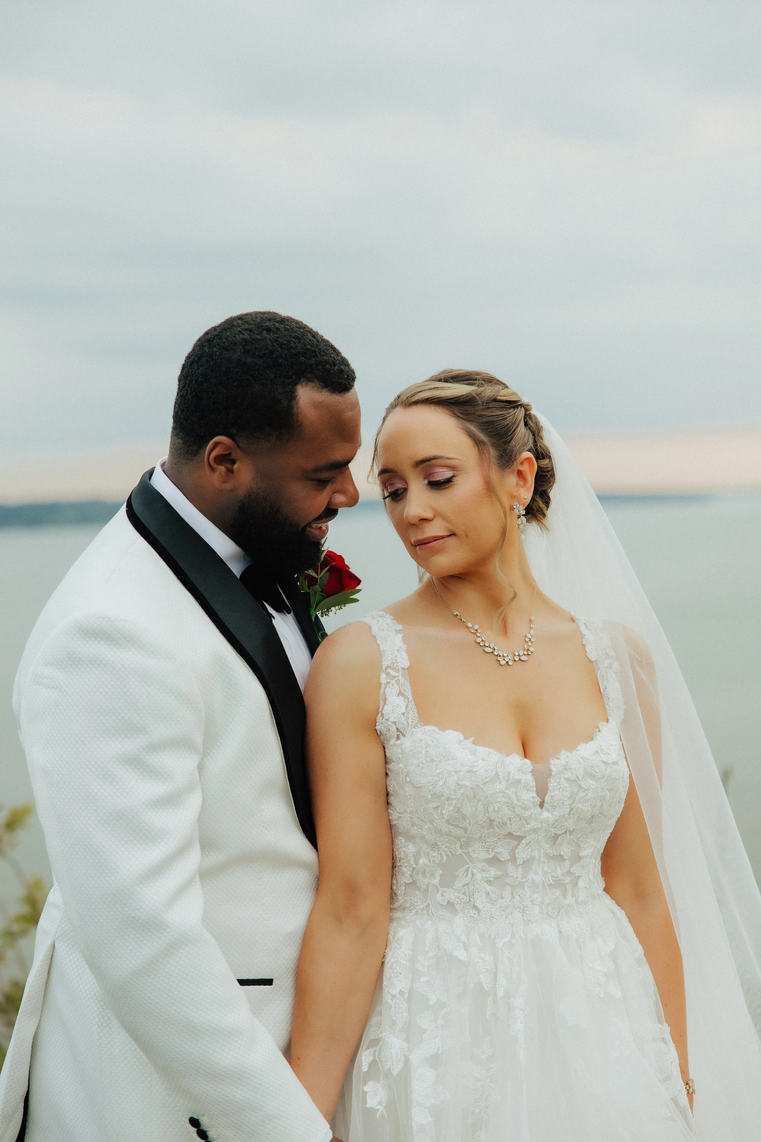 A bride and groom are standing close together outdoors, with the groom holding the bride's hand. The bride is wearing a white wedding dress with lace details and a pearl necklace, and has her hair styled with a braid. The groom is in a white tuxedo w