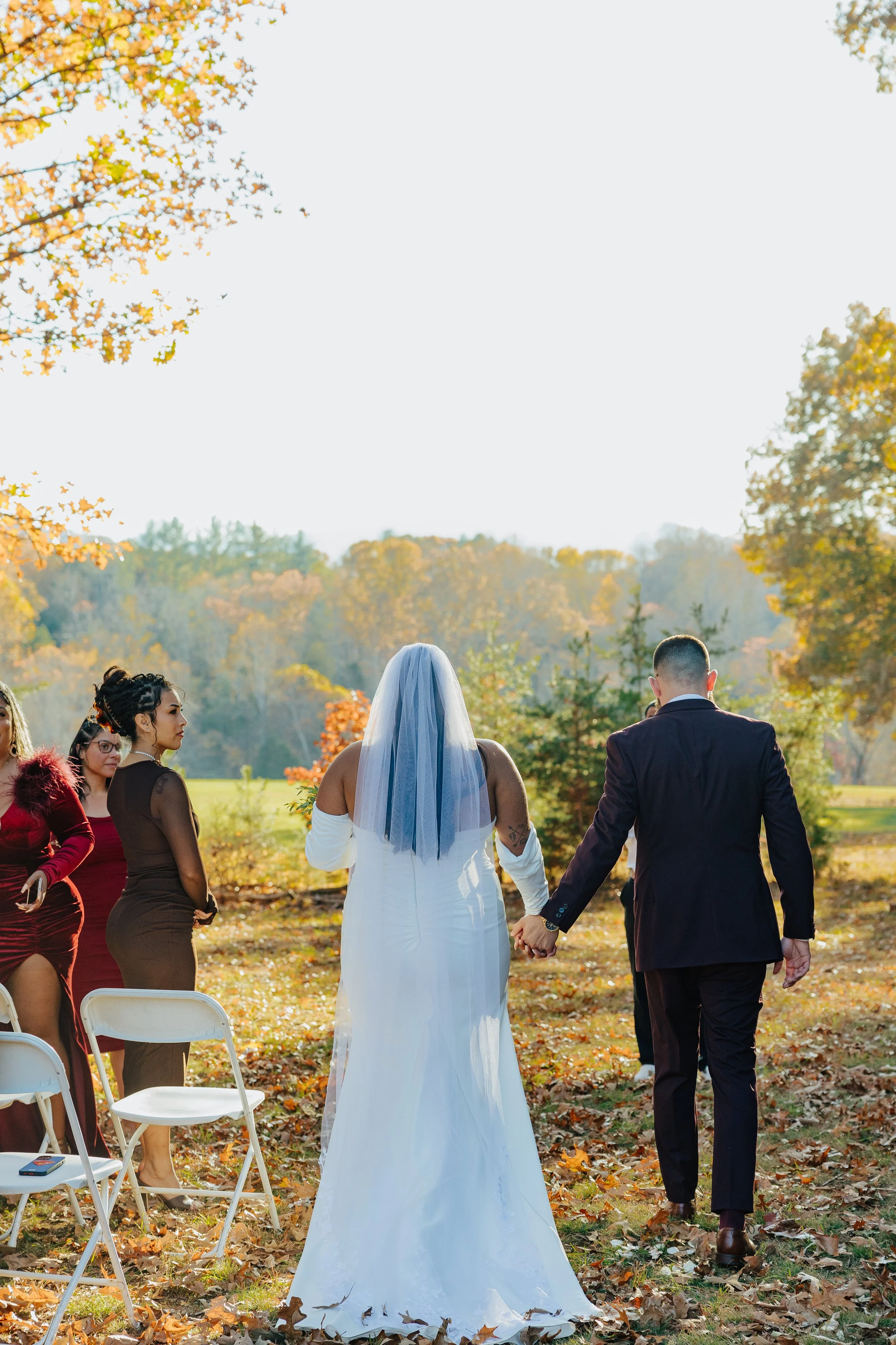 A couple dressed in wedding attire holding hands and walking away from the camera during an outdoor wedding ceremony in fall, with guests seated nearby.