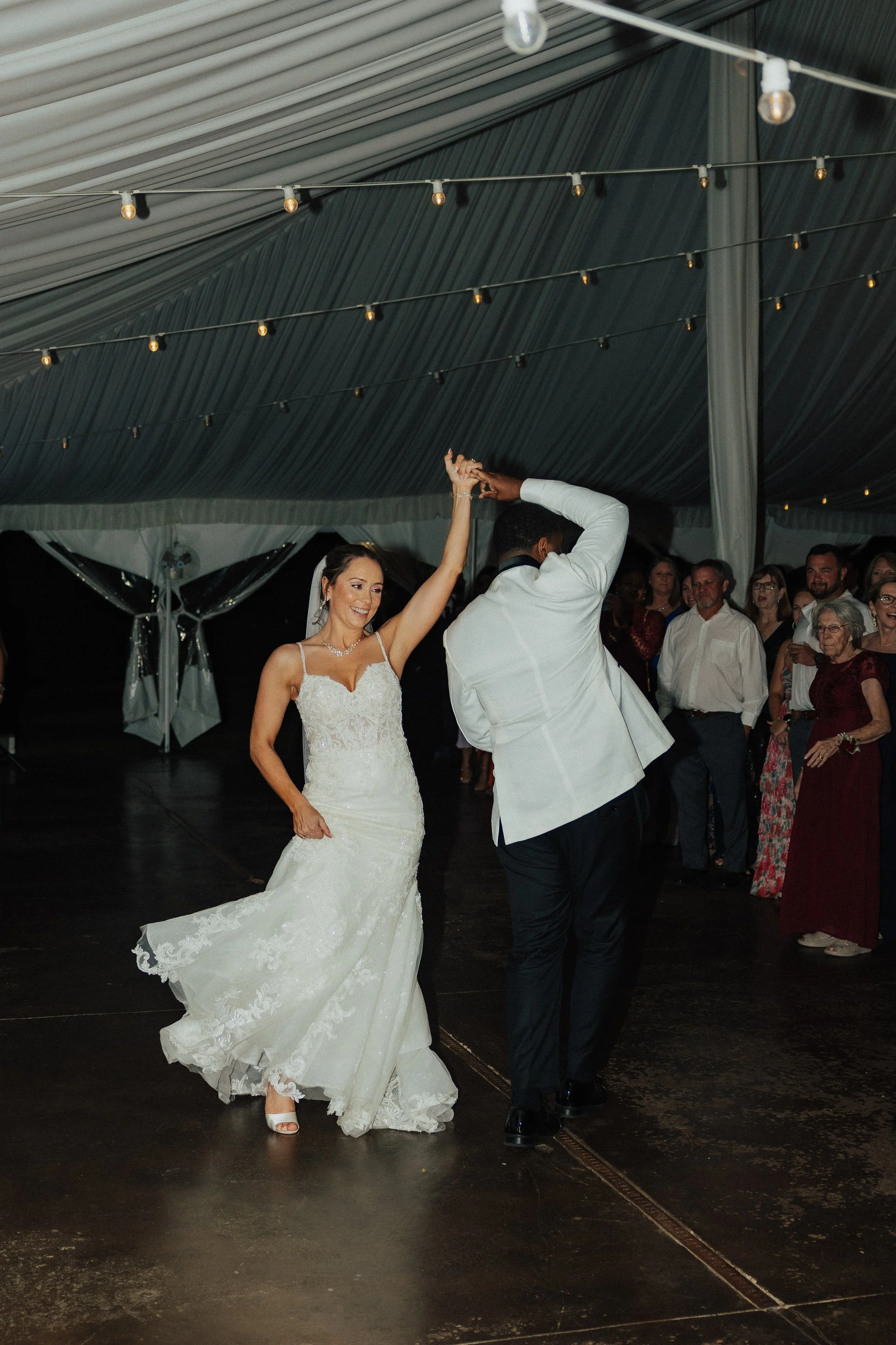 A bride and groom dancing at their wedding reception in a decorated tent with string lights and onlookers in the background.