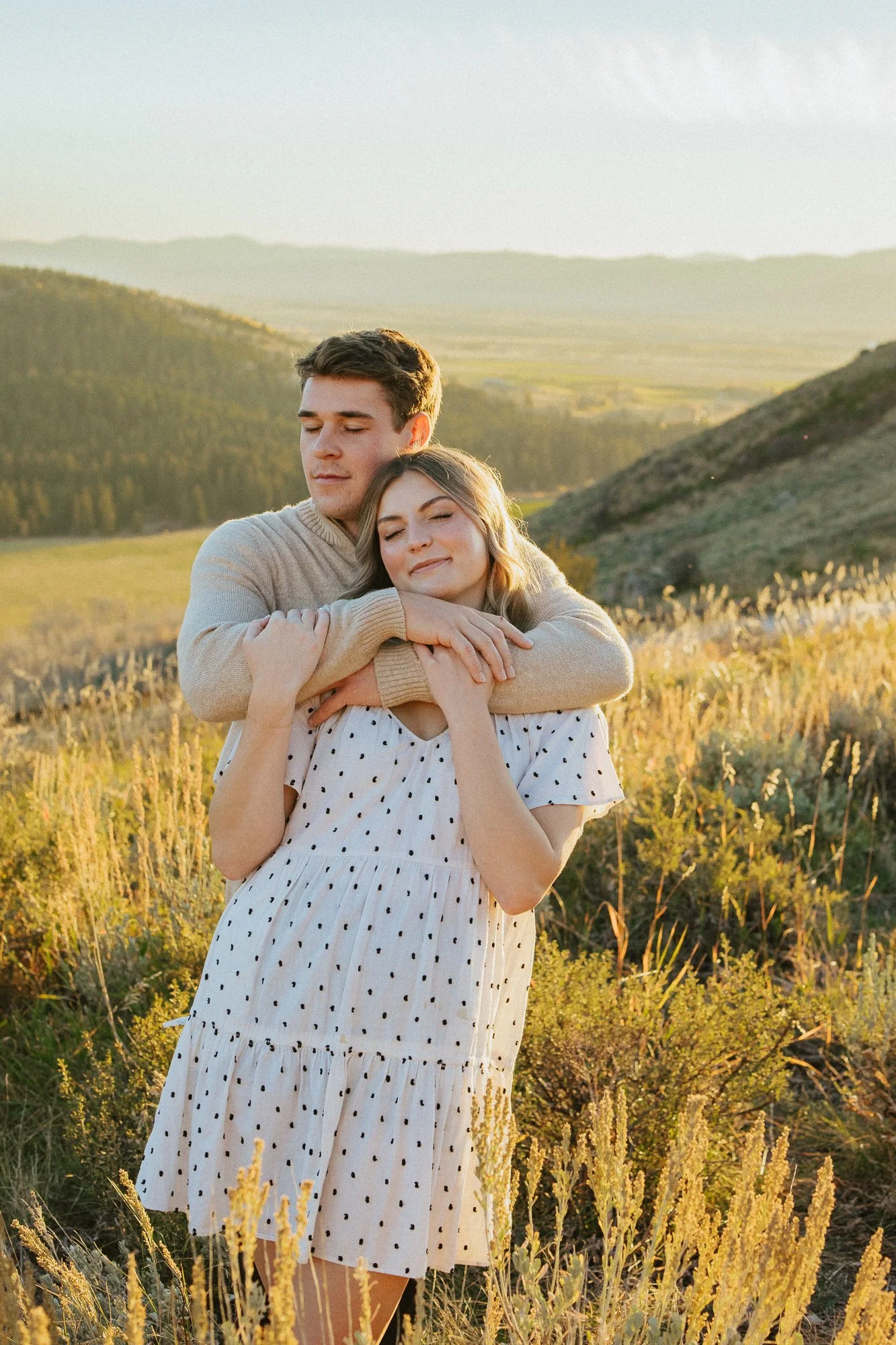 A young man and woman embracing in a field of yellow flowers with rolling hills in the background during sunset, both with their eyes closed, conveying a sense of tranquility and affection.