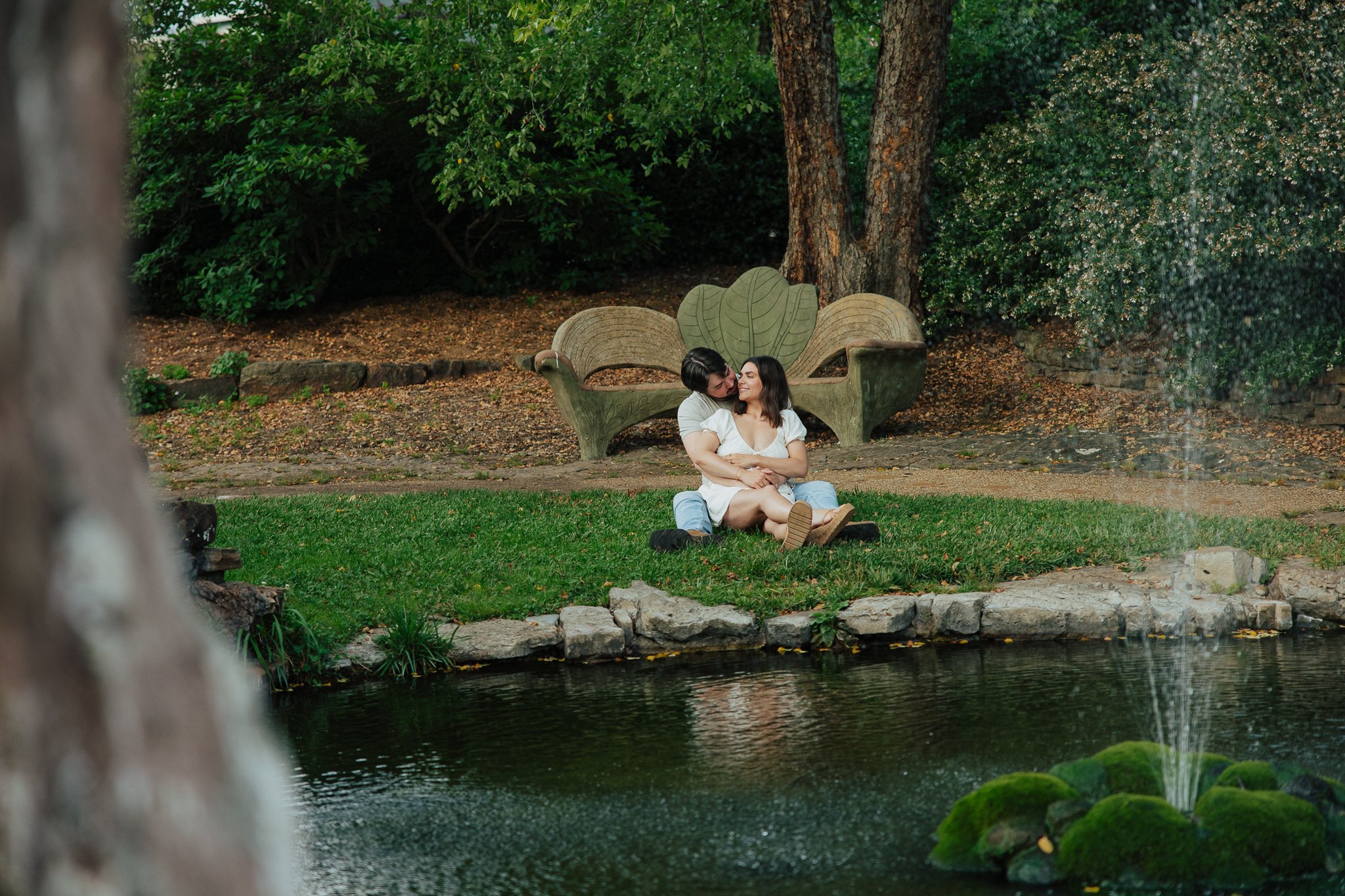 A couple sitting on grass by a pond in a park, hugging each other, with trees and bushes in the background.