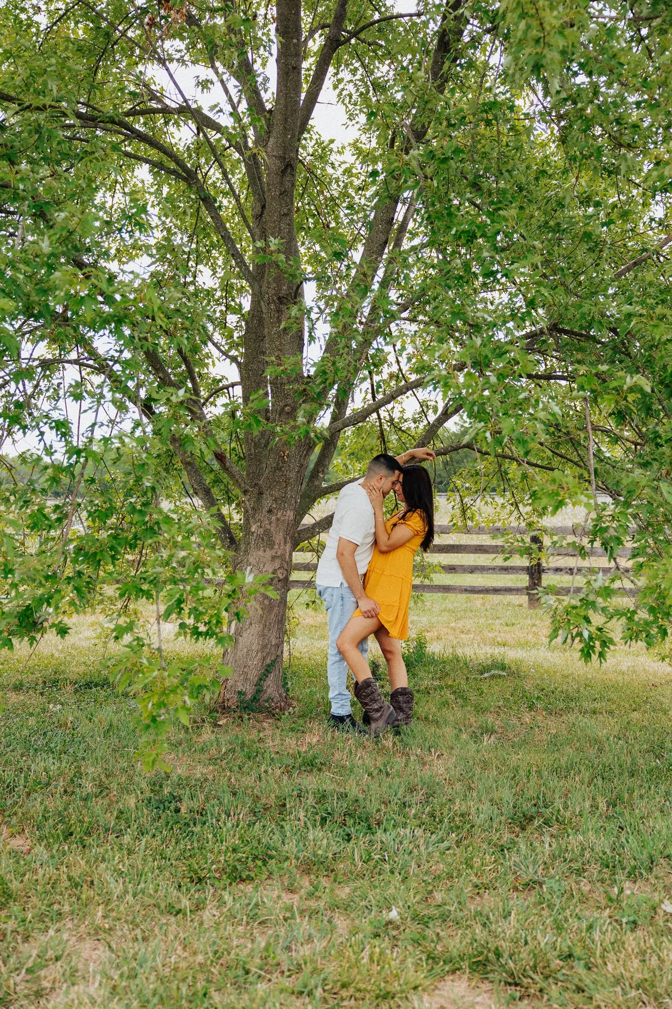 Couple standing under a large tree in a rural setting, embracing and looking at each other, with a wooden fence in the background and green grass on the ground.