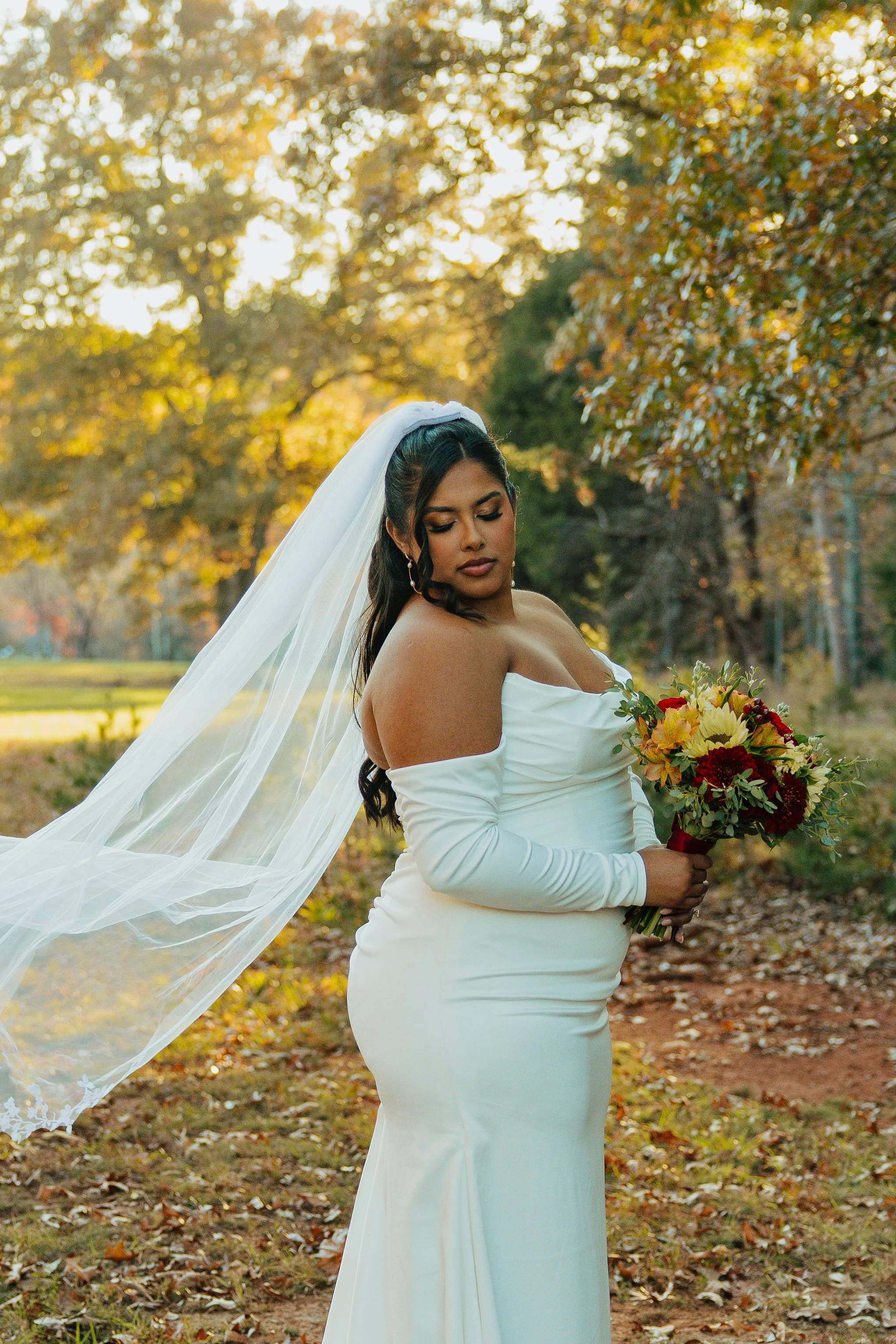 A woman in a white wedding dress with a veil holding a bouquet of yellow and red flowers outdoors during autumn with trees and fallen leaves in the background.