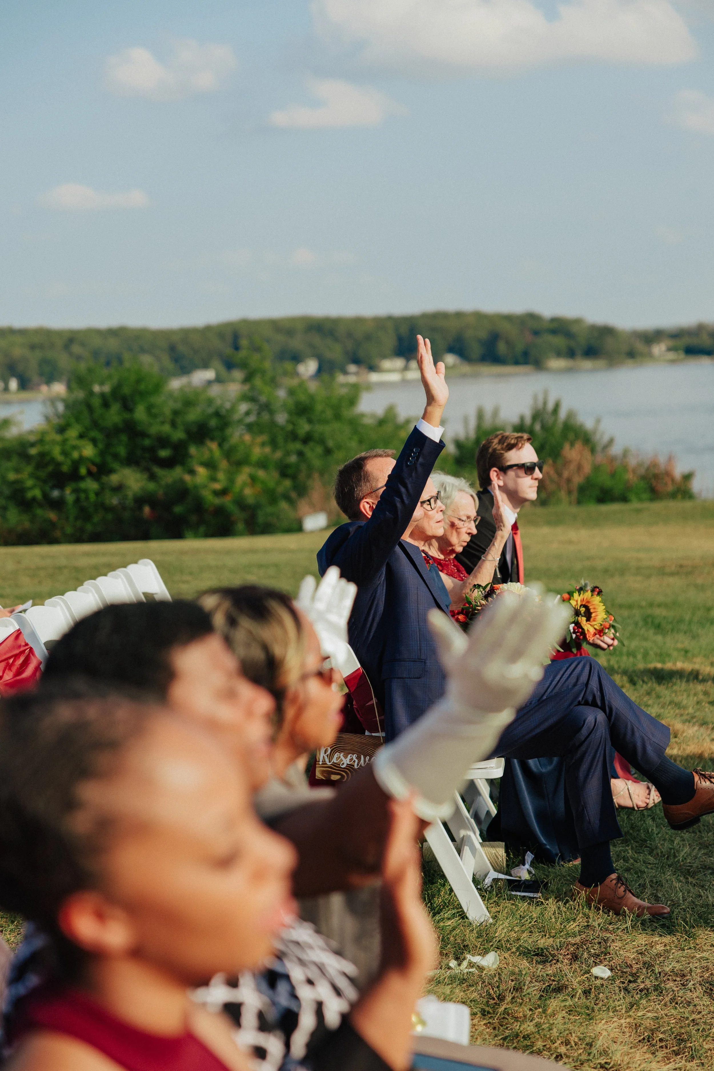 People seated outdoors on white chairs attending a formal event near a lake, with some individuals raising their hands, including a man in a navy suit and sunglasses, and elderly women holding bouquets, with trees and water in the background.