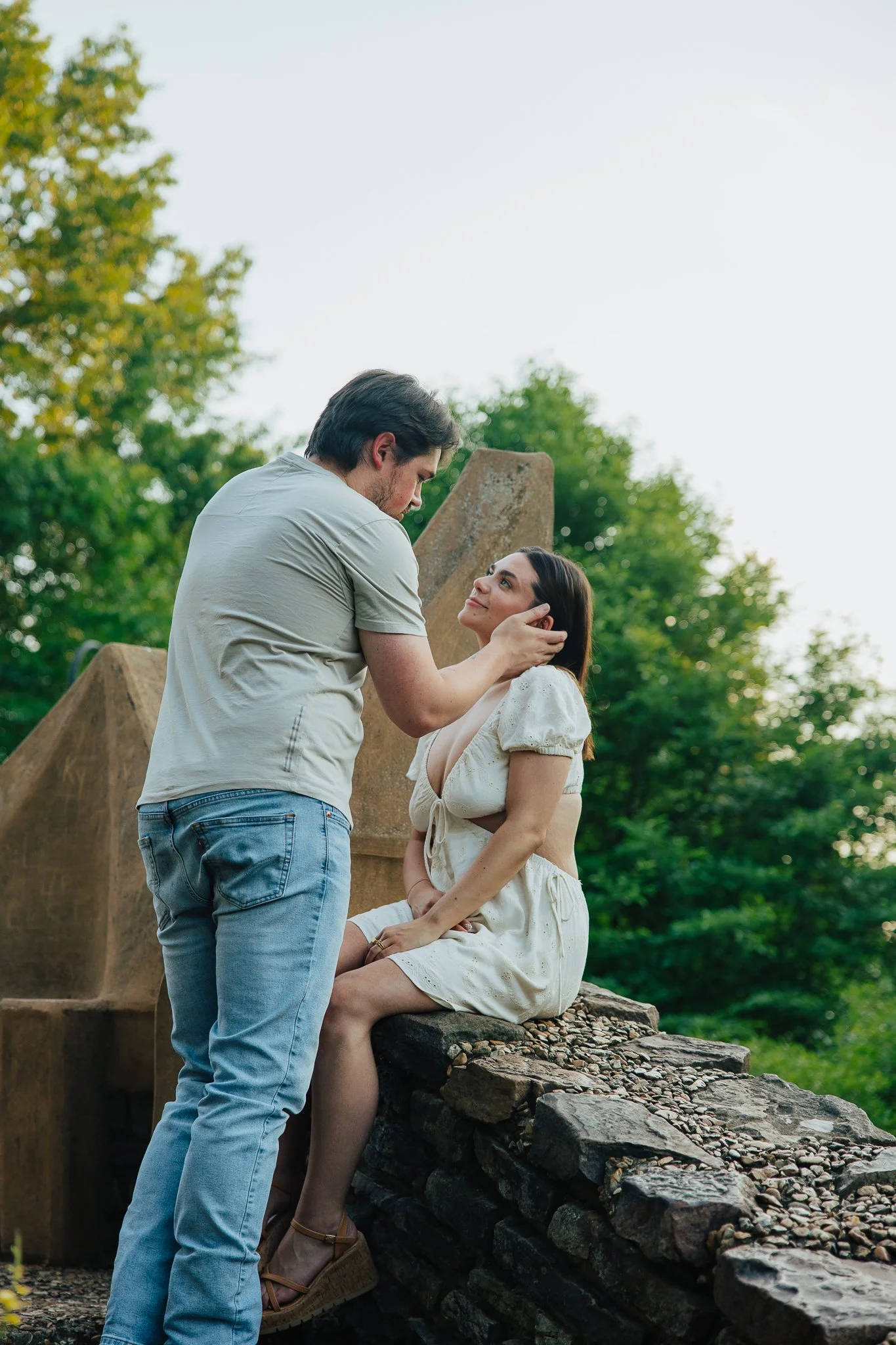 A man and woman are outside in a romantic moment, with the man gently holding the woman's face as she sits on a stone ledge, surrounded by green trees and clear sky.