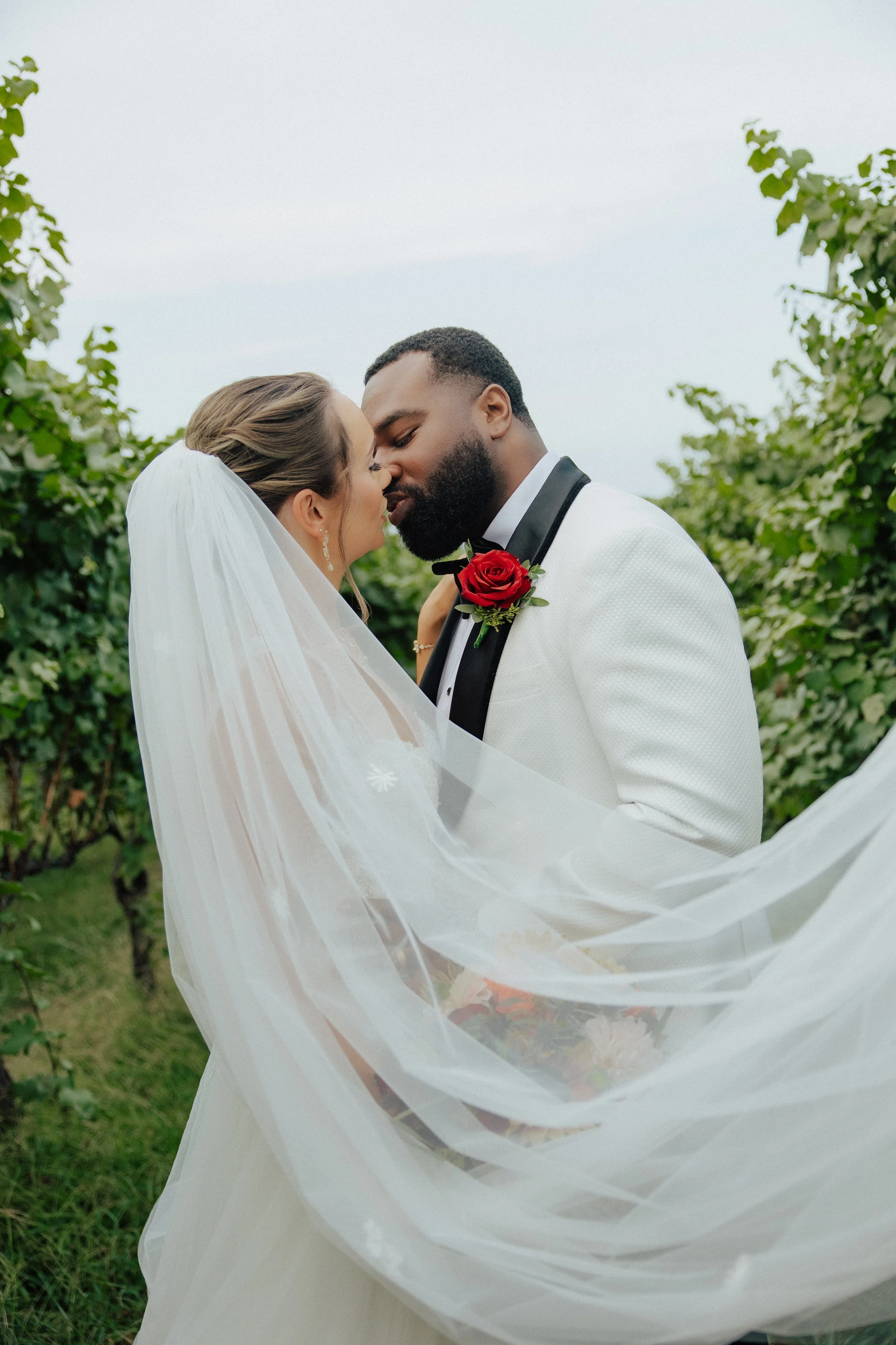 A newlywed couple is sharing a romantic moment outdoors in a garden, with the bride wearing a white veil and dress, and the groom in a white tuxedo with a black lapel and a red rose boutonniere, with greenery in the background.