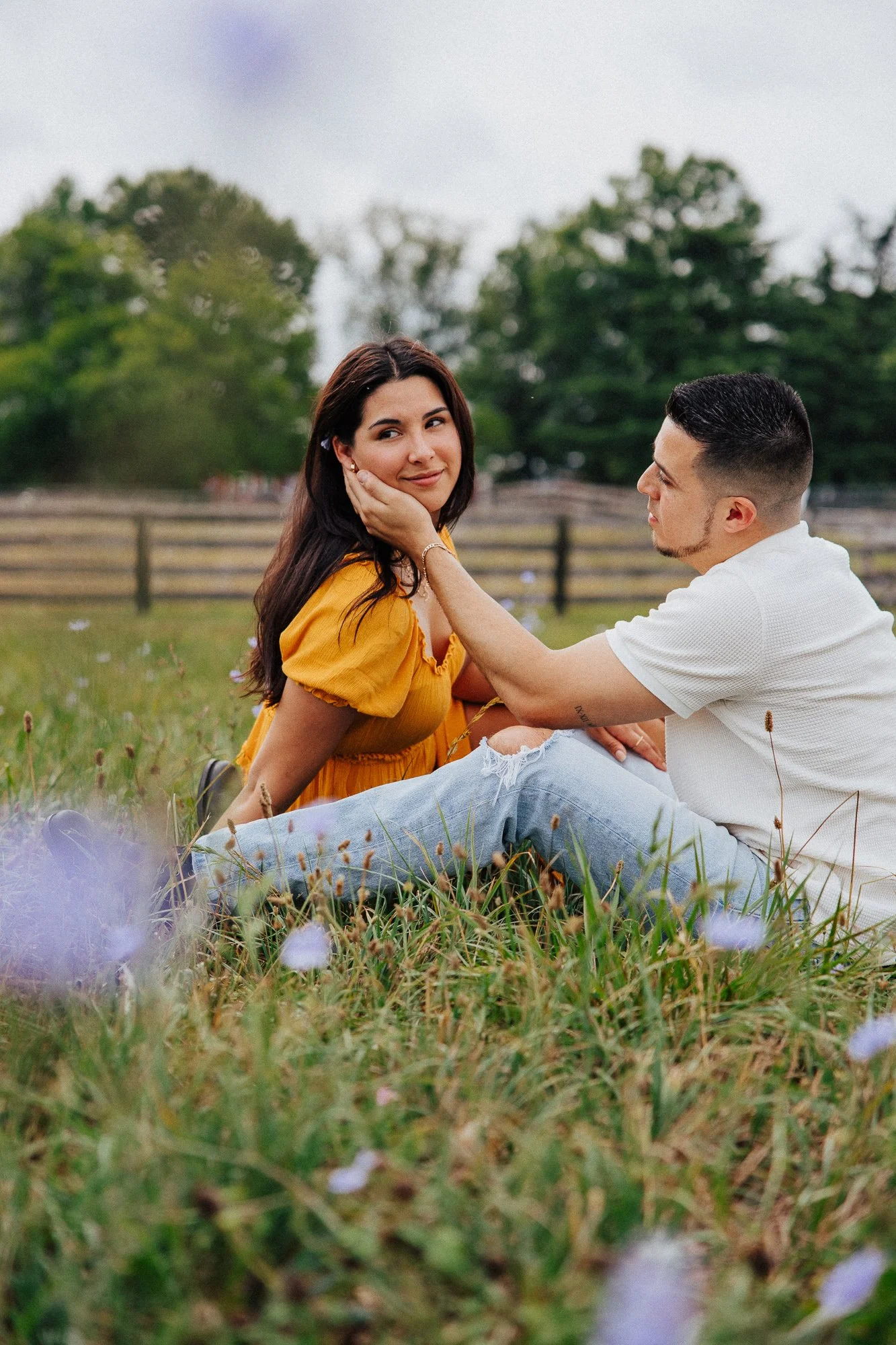 A man and woman sitting on grass in a park, the woman wearing a yellow dress and the man in a white shirt, with trees and a fence in the background.
