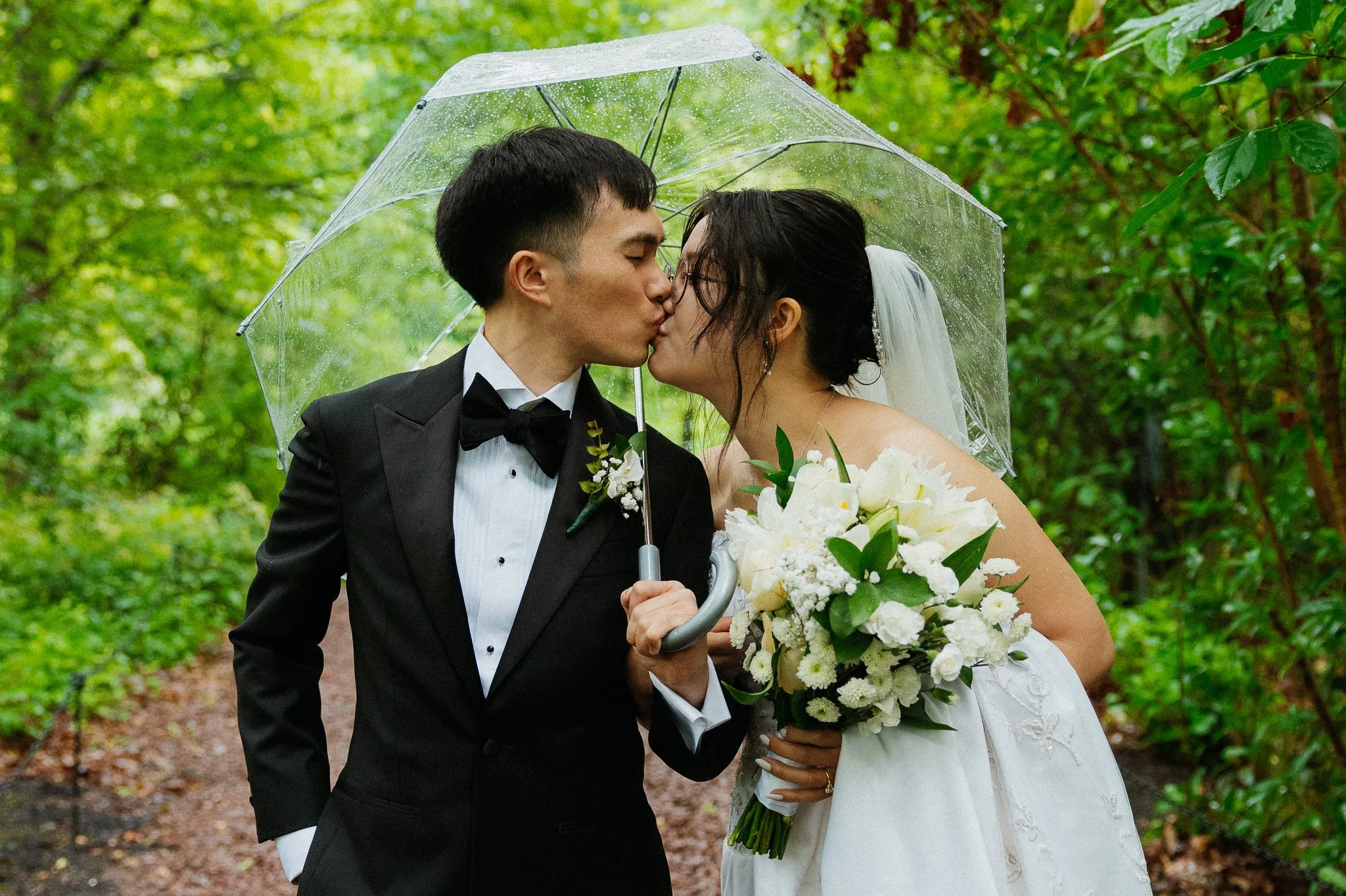 A bride and groom kiss under a clear umbrella in a green outdoor setting, with the bride holding a bouquet of white flowers.