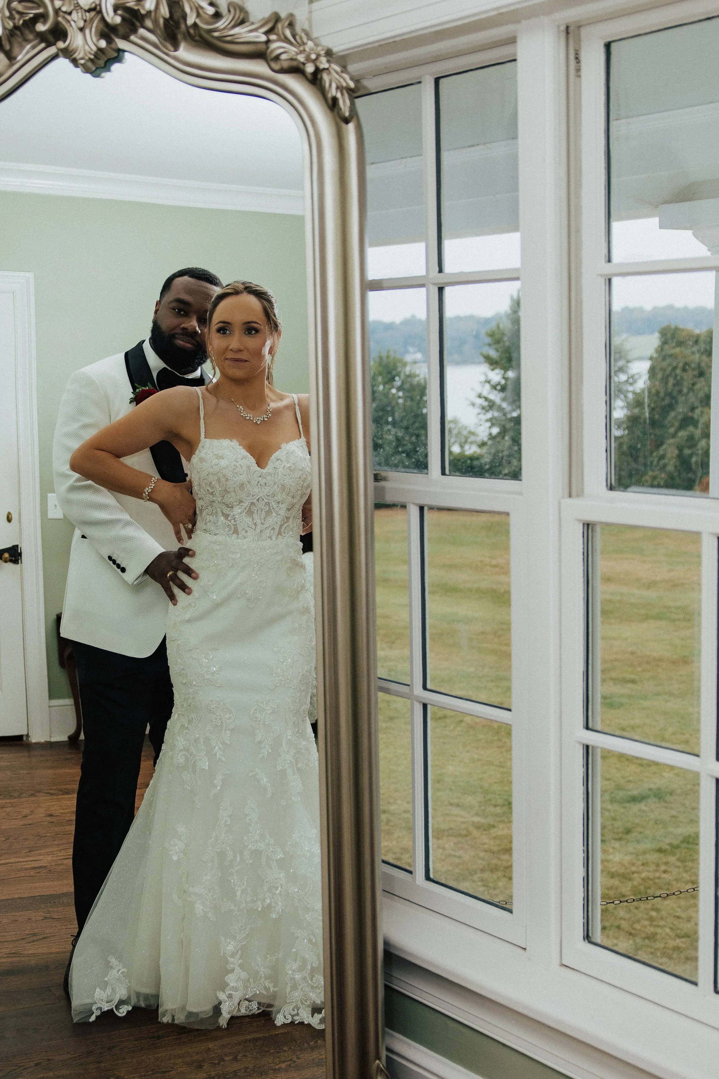 A couple in wedding attire taking a mirror selfie inside a room with large windows overlooking a grassy yard and water.